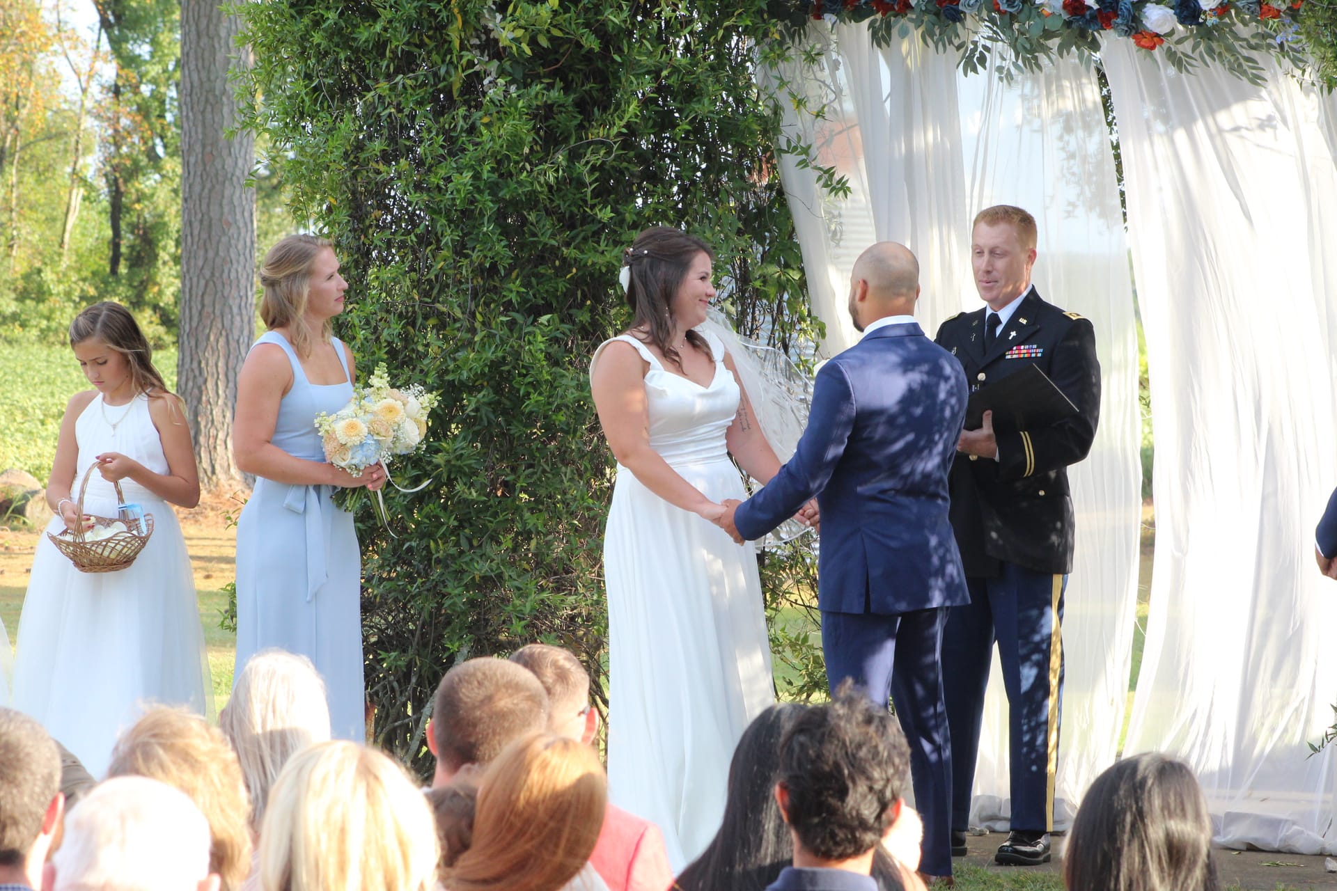 Bride in white gown holds hands with groom in navy suit during outdoor wedding ceremony officiated by a military officer beneath a draped floral arch at Scarborough House.
