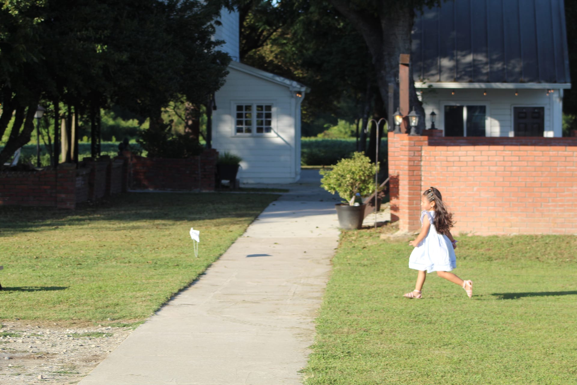 A young flower girl in a white dress runs across the lush green lawn in front of the historic white manor house at Scarborough House wedding venue on a sunny day.