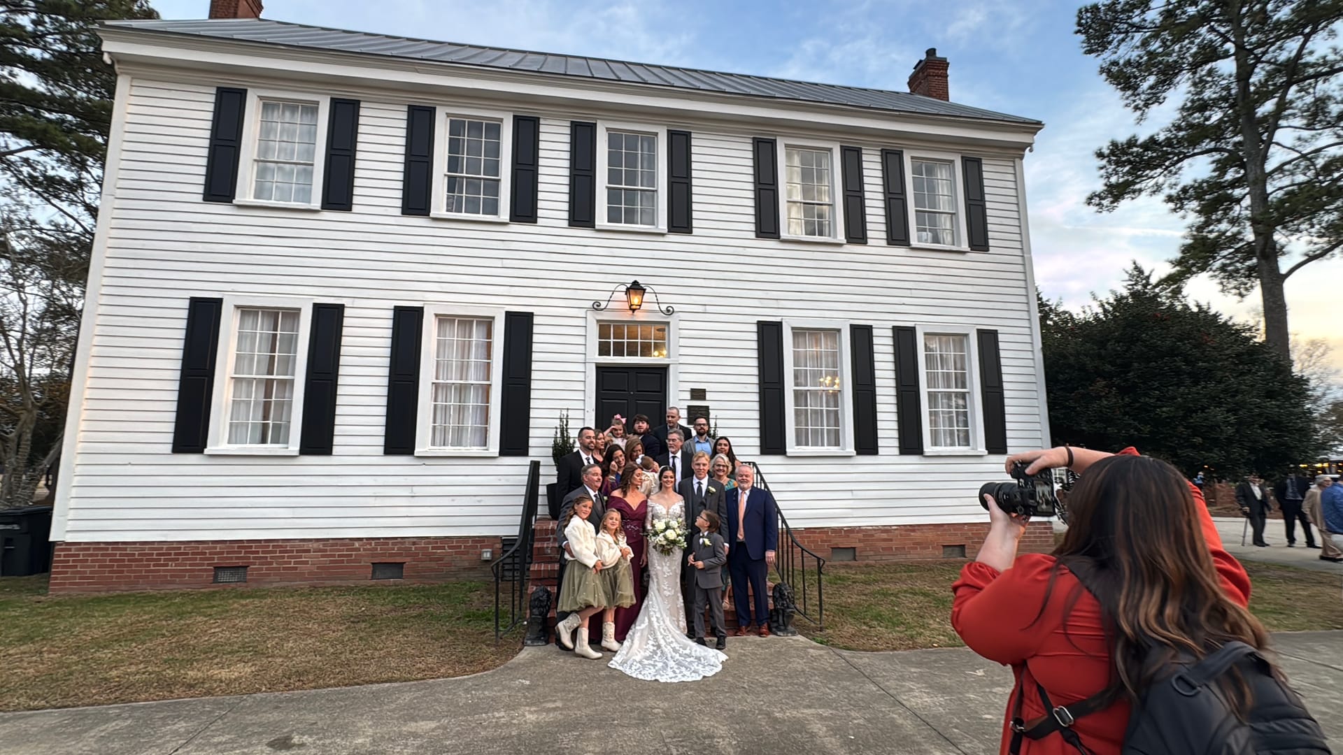 Bride and groom pose with their full wedding party and family on the front steps of the 1821 Scarborough House white clapboard manor while a photographer in red captures the moment.