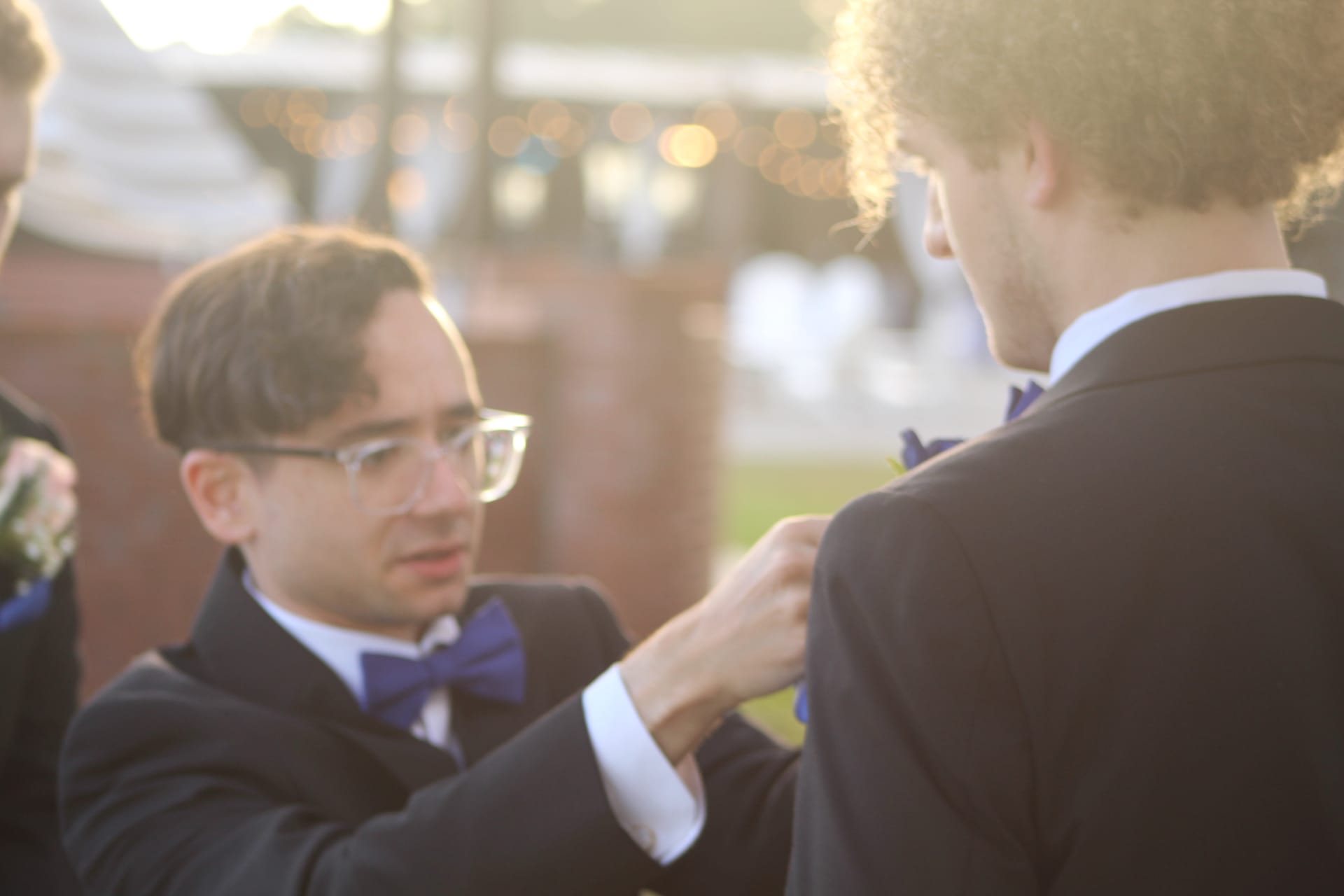 A groomsman in a black suit and navy bow tie adjusts a boutonniere on another groomsman outdoors in warm golden evening light.