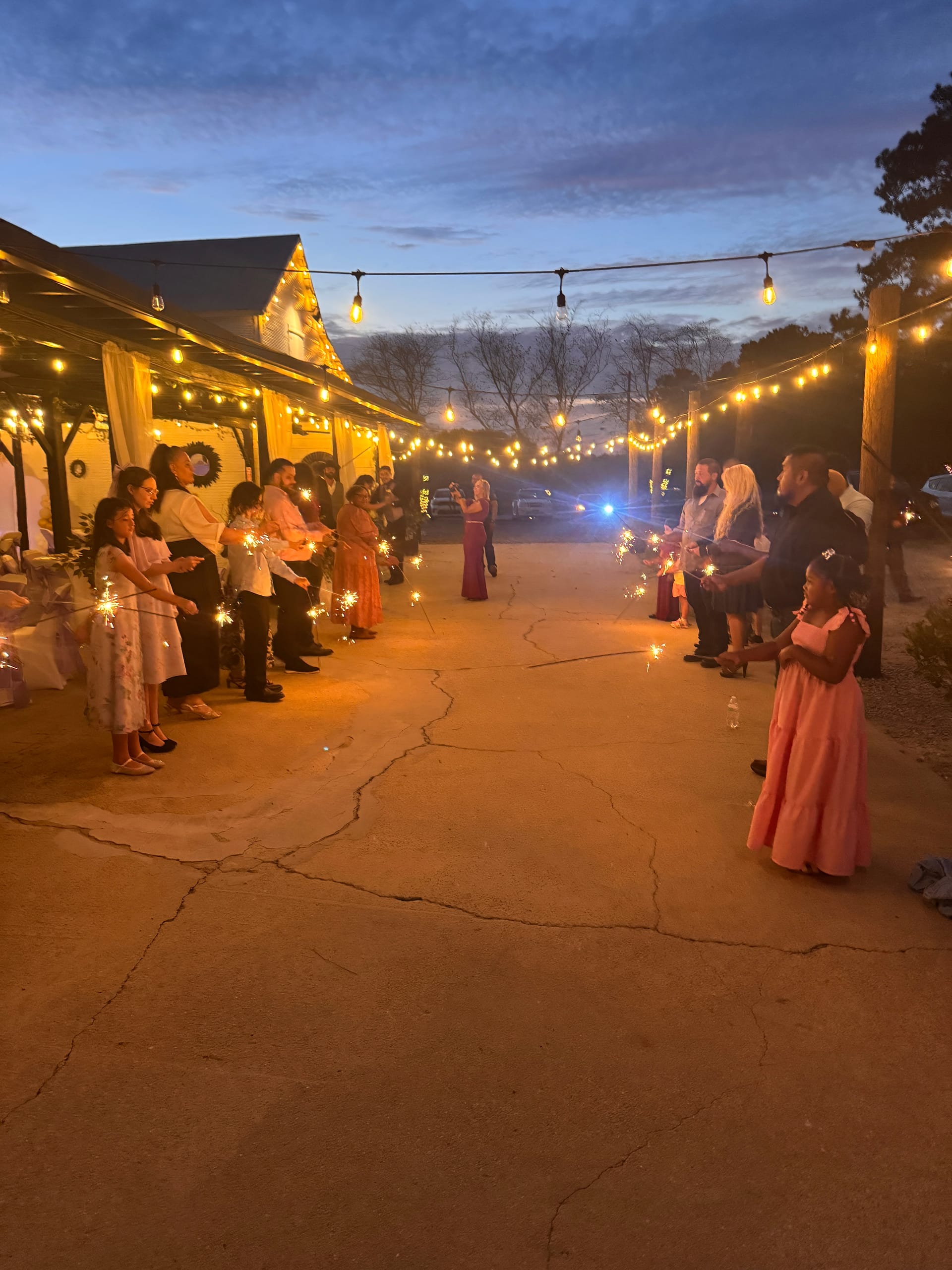 Wedding guests holding sparkling sparklers and forming a send-off tunnel under warm string lights at dusk outside a historic estate venue.