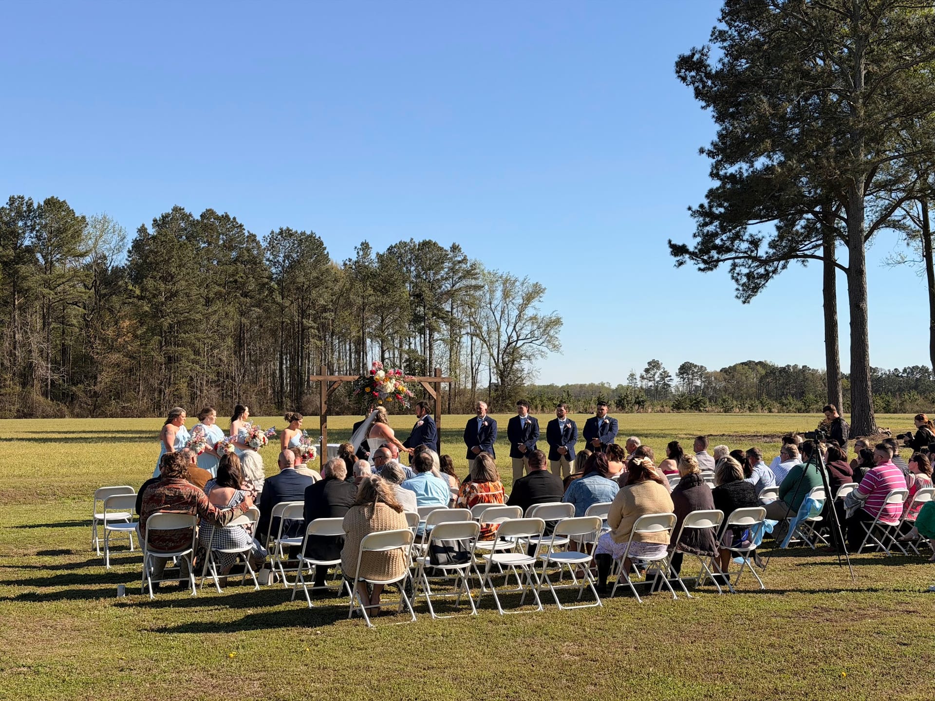 Outdoor wedding ceremony on a sunny day at a rural estate with guests seated in white folding chairs facing a rustic wooden arch adorned with colorful florals and the wedding party standing before a backdrop of open farmland and pine trees.