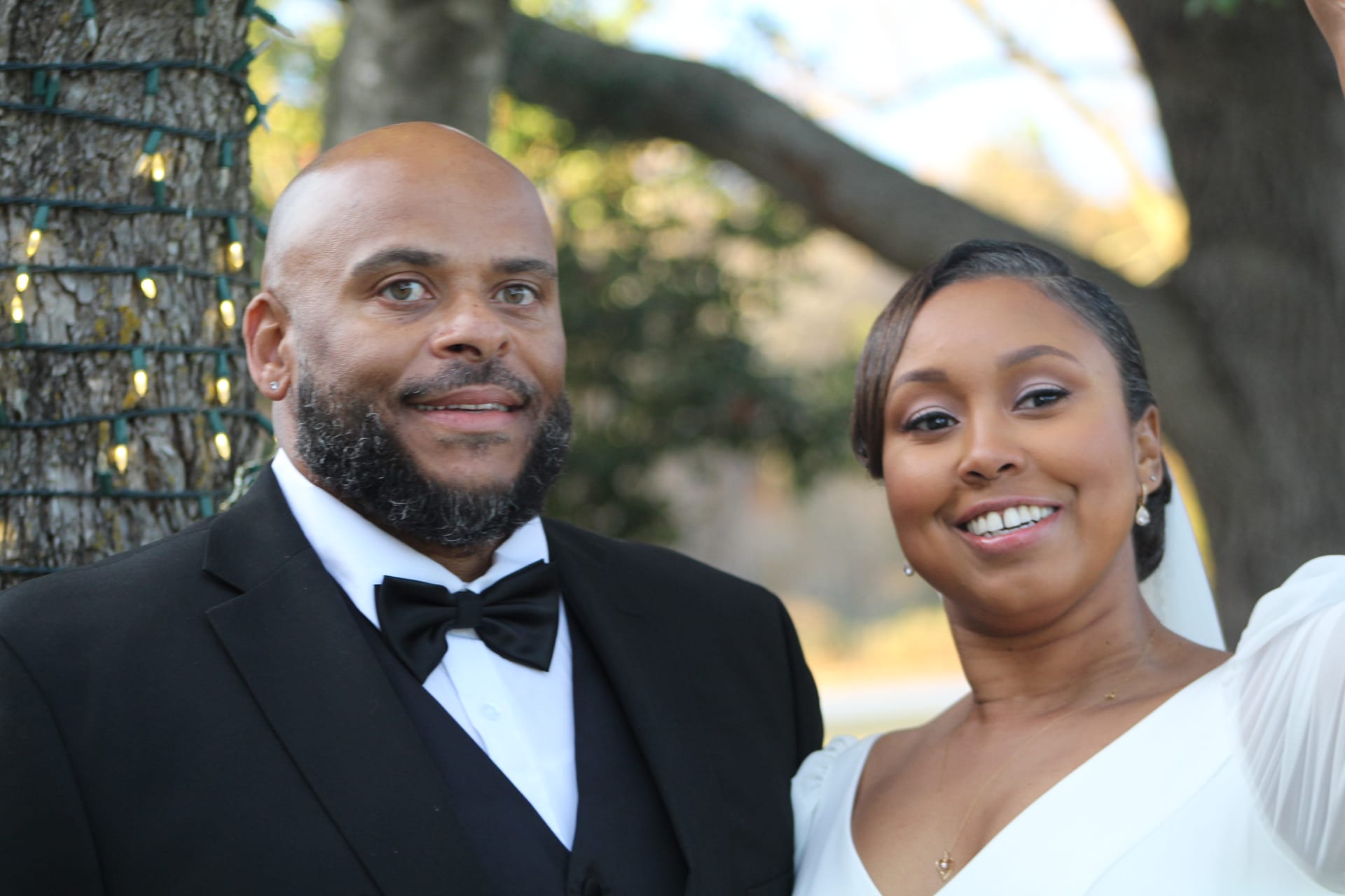 Bride in a white gown and groom in a black tuxedo with bow tie pose together smiling in front of a large oak tree adorned with string lights at Scarborough House.