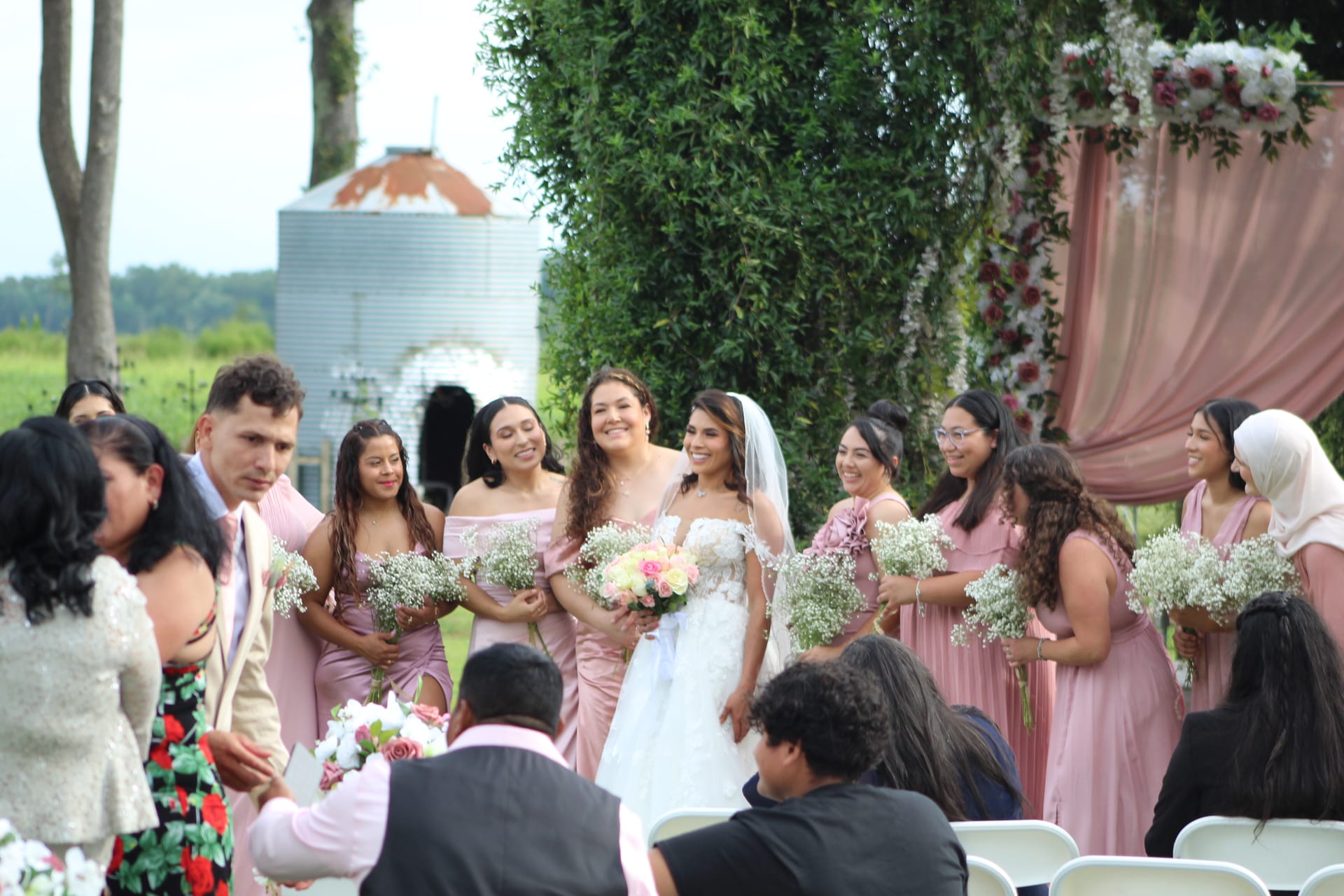A radiant bride in a white gown and veil walks alongside her bridesmaids in blush pink dresses holding baby's breath bouquets toward a flower-adorned ceremony arch on the estate lawn.
