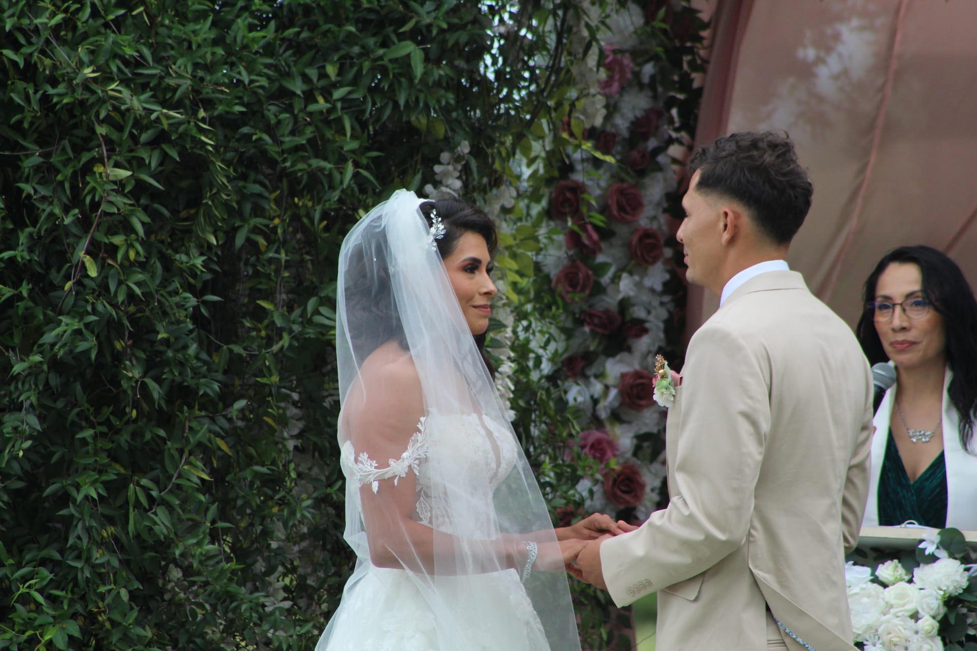 Bride and groom hold hands and exchange vows outdoors at a garden ceremony framed by lush greenery and a floral rose backdrop while the officiant looks on.