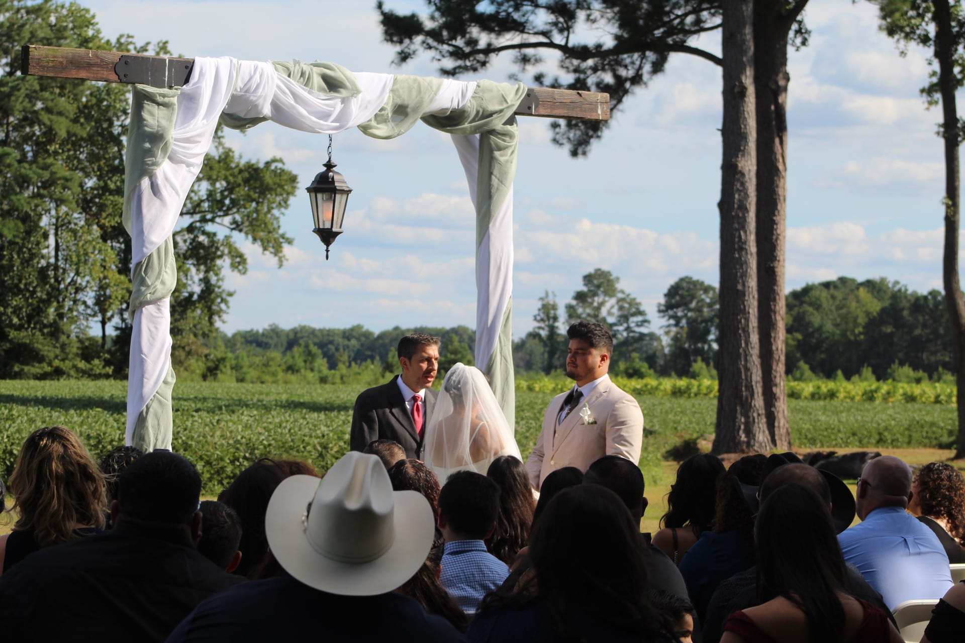 Outdoor wedding ceremony under a rustic wooden arch draped in white and sage fabric with a hanging lantern, groom and officiant standing with veiled bride before a lush green farmland backdrop at Scarborough House.