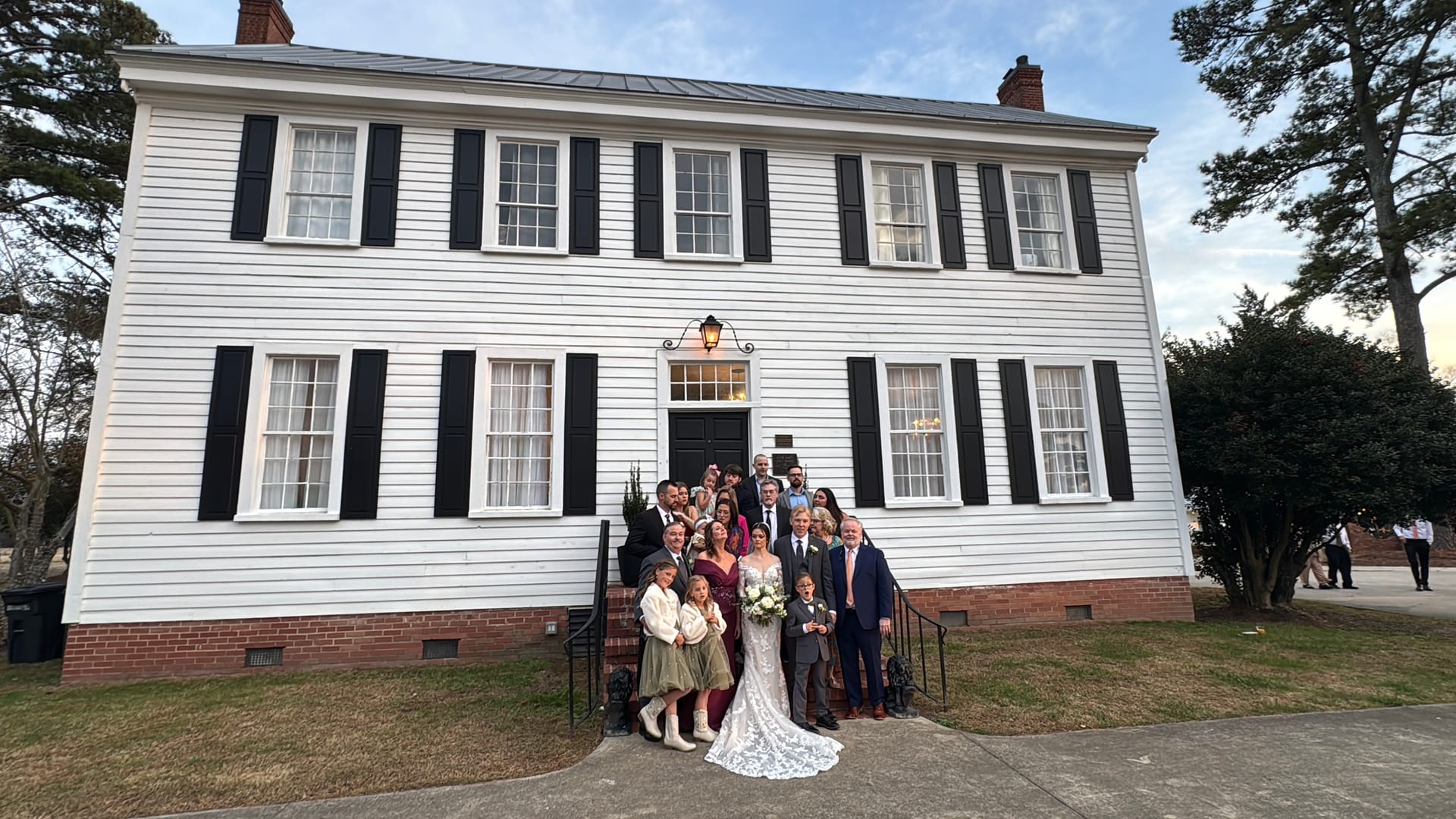 Bride and groom surrounded by their wedding party and family on the front steps of the historic 1821 Scarborough House manor at dusk.
