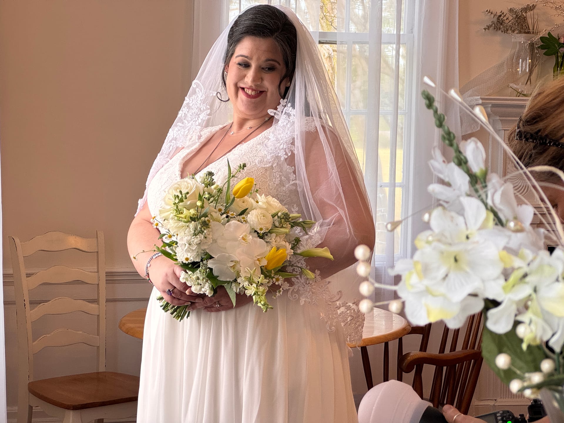 A radiant bride in a lace wedding gown and cathedral veil smiles softly while holding a bouquet of white roses and yellow tulips inside a warmly lit historic estate room.