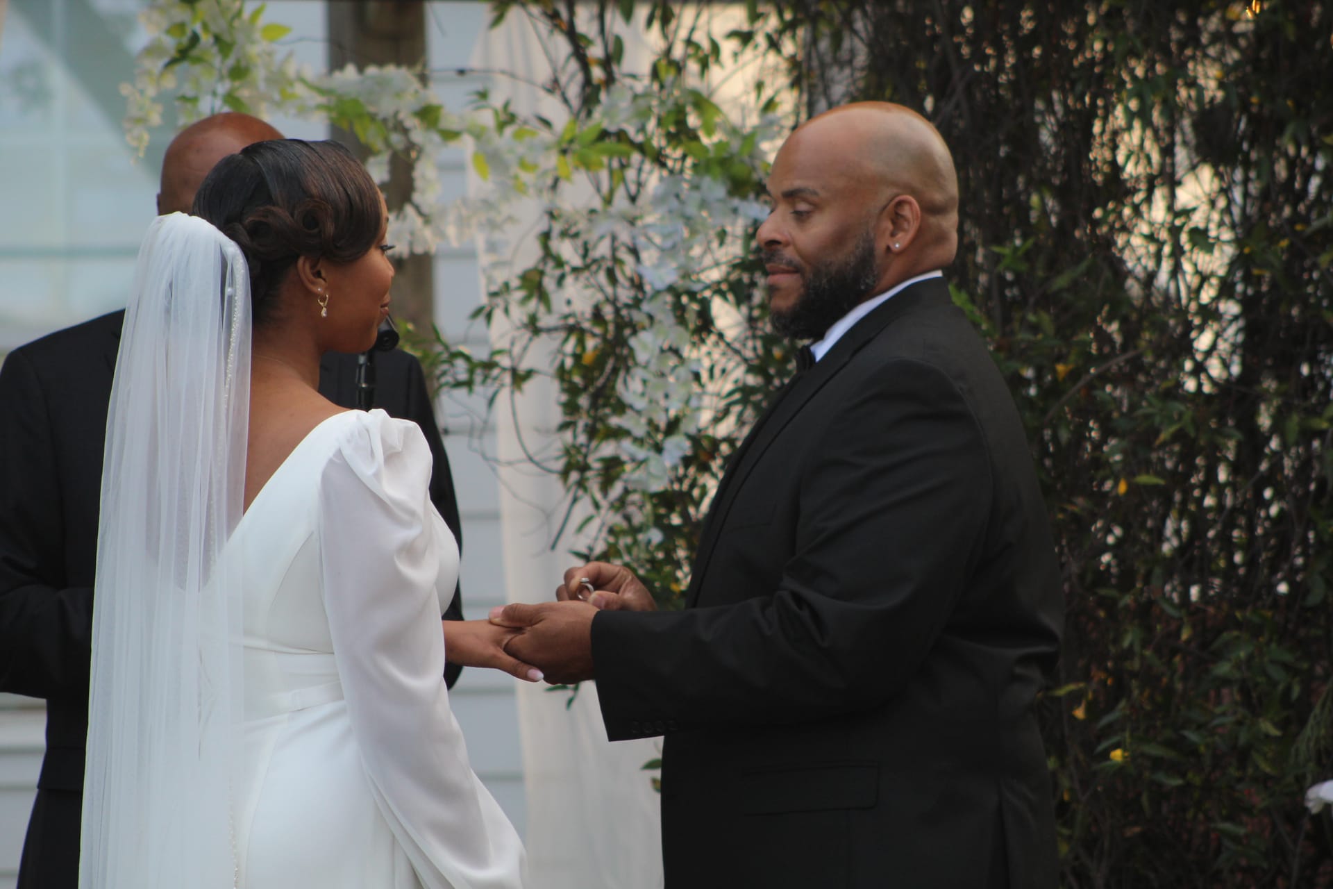 Groom in a black tuxedo places a ring on his bride's finger during an outdoor wedding ceremony at Scarborough House, surrounded by lush greenery and white floral arrangements.