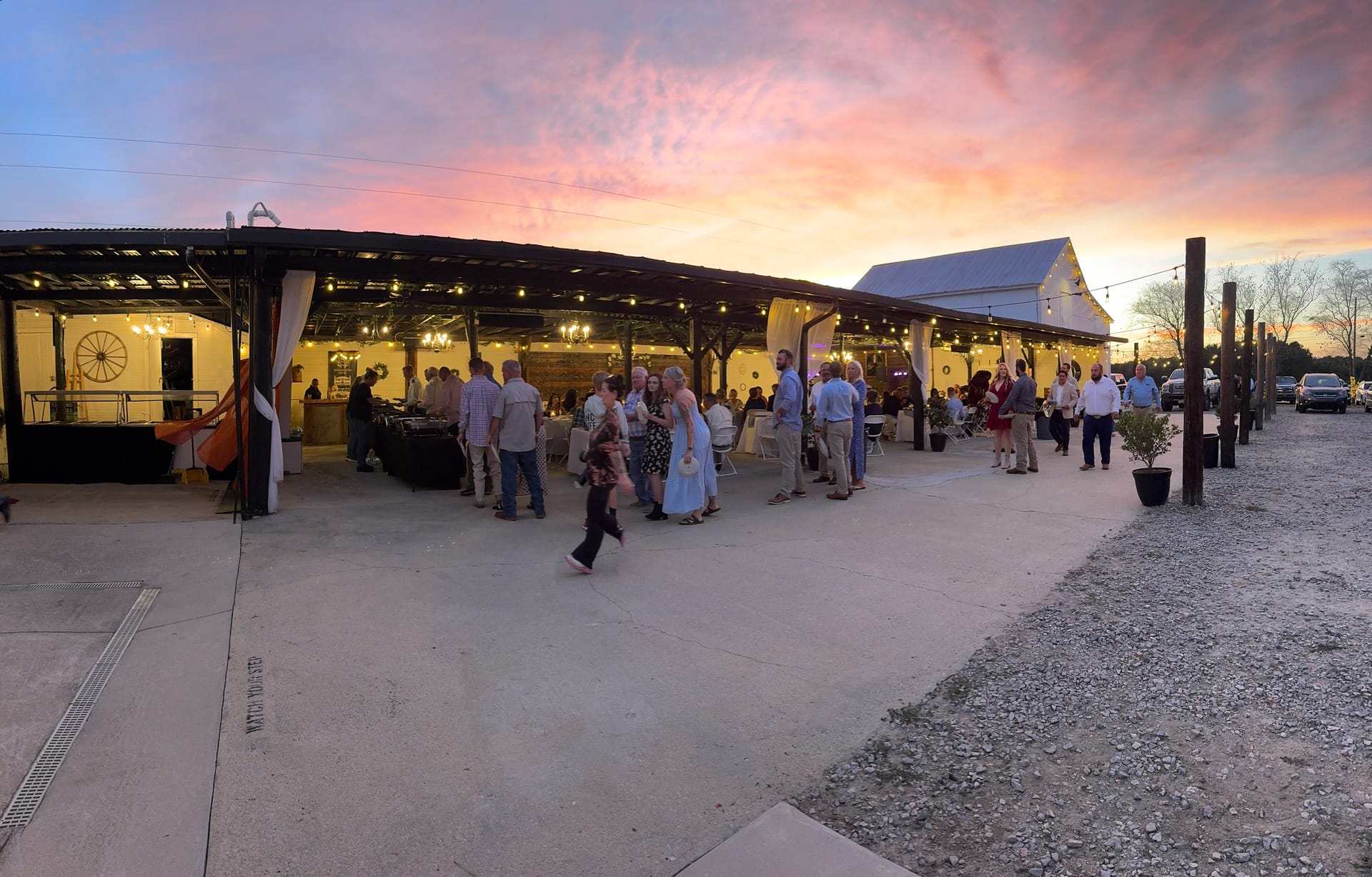 Wedding guests mingle under a warmly lit open-air pavilion at Scarborough House as a dramatic pink and orange sunset fills the sky behind the historic white barn.