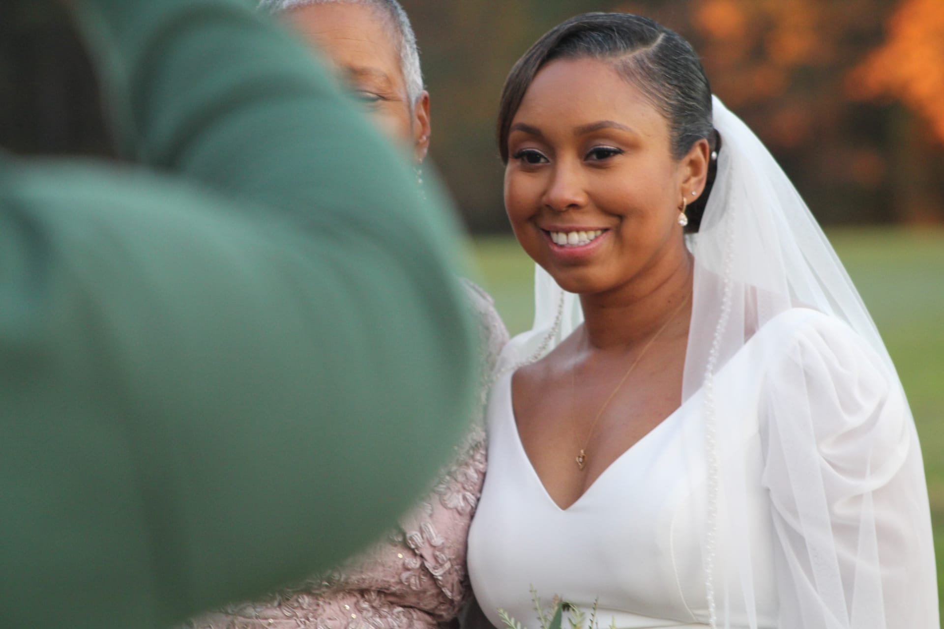 A glowing bride in a white gown and veil smiles warmly during an outdoor ceremony at Scarborough House estate in soft golden-hour light.