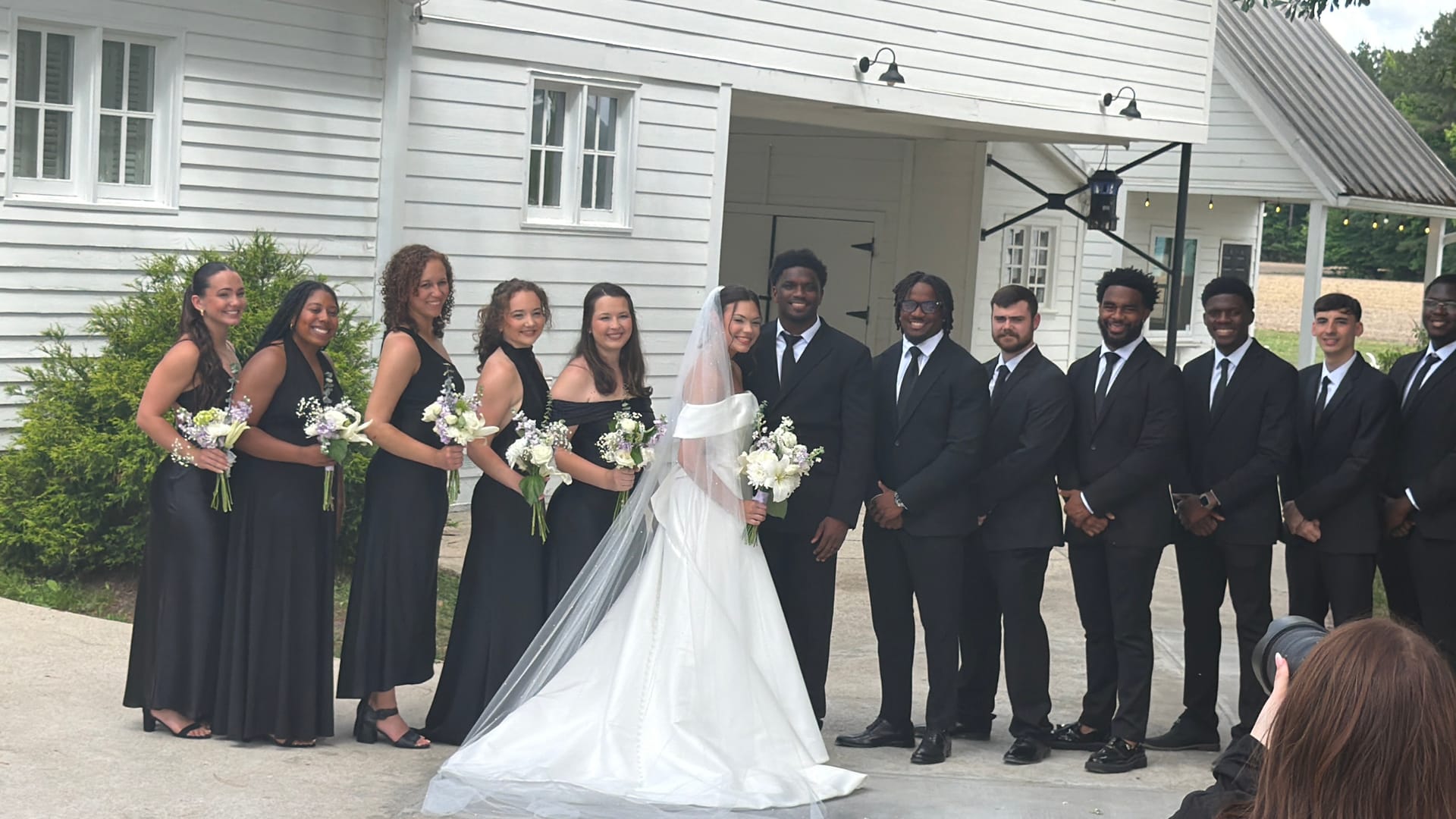Wedding party in coordinated black dresses and suits poses with the bride in a flowing white gown and veil in front of the white historic Scarborough House venue.