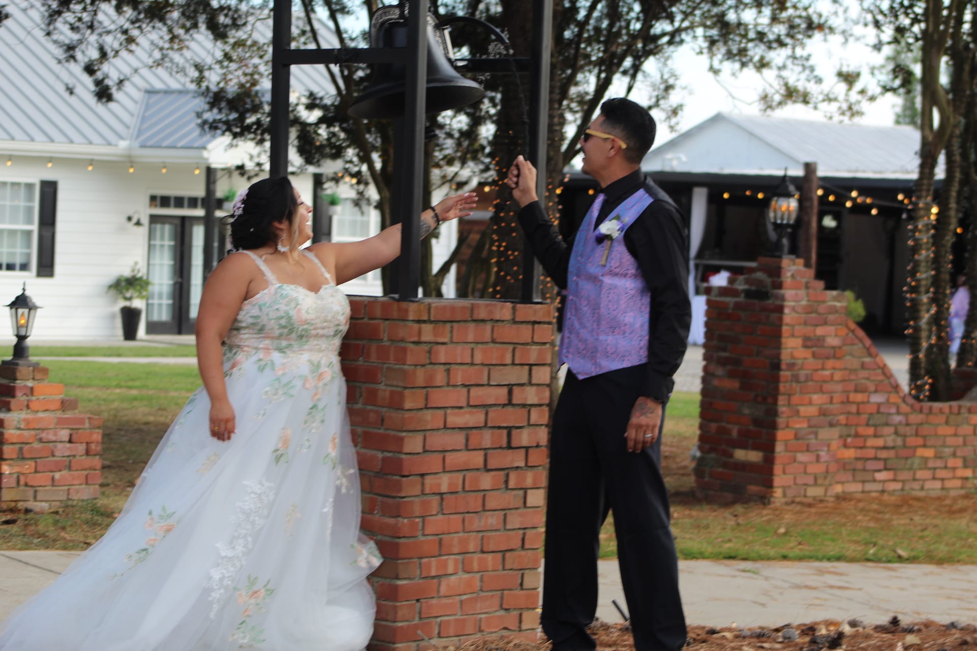 Bride in a floral ballgown and groom in a purple vest reach up together to ring the decorative bell at Scarborough House wedding venue.