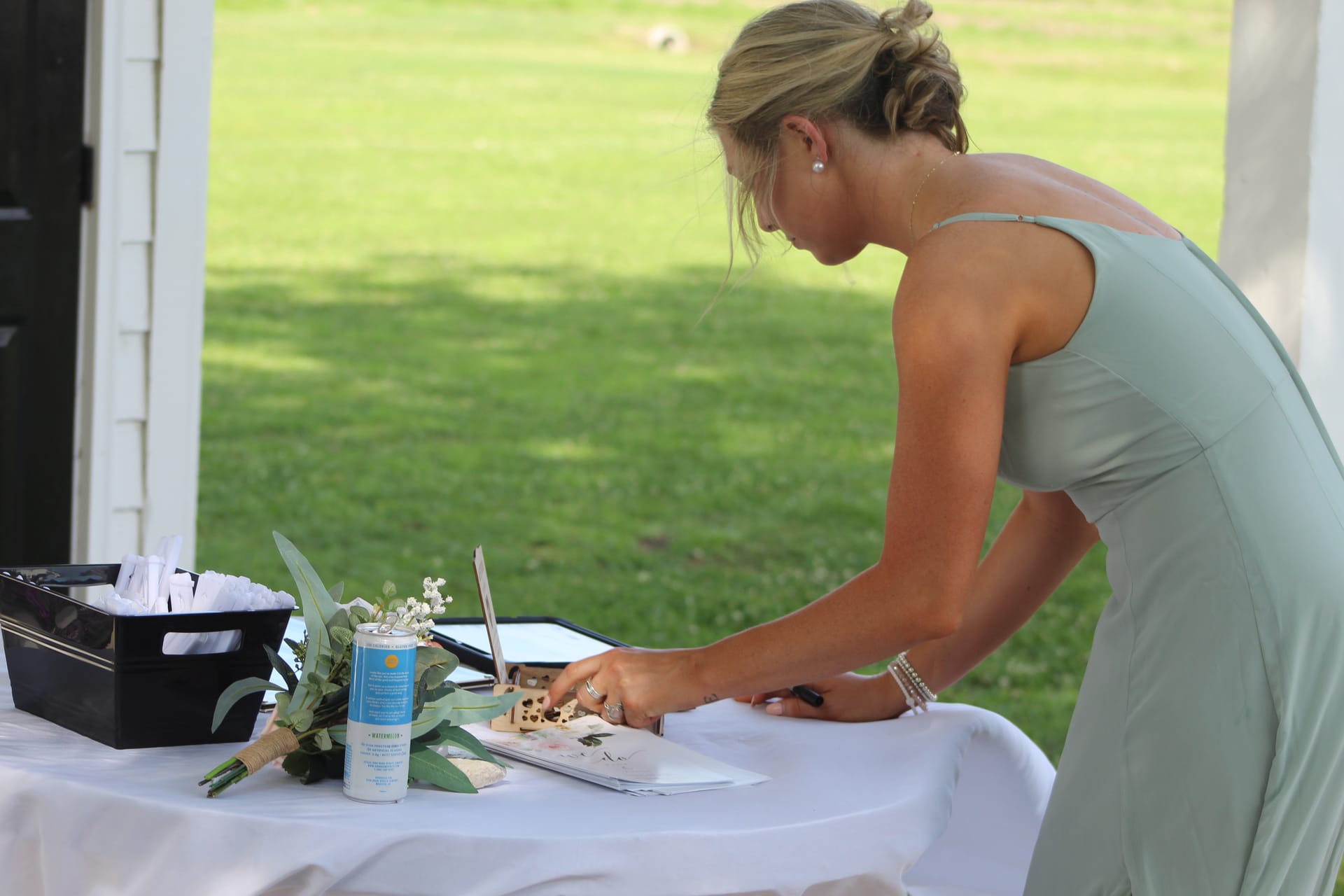 A bridesmaid in a sage green gown leans over a white-draped table to sign the wedding guest book beneath the covered porch of Scarborough House, with lush green lawns visible in the background.
