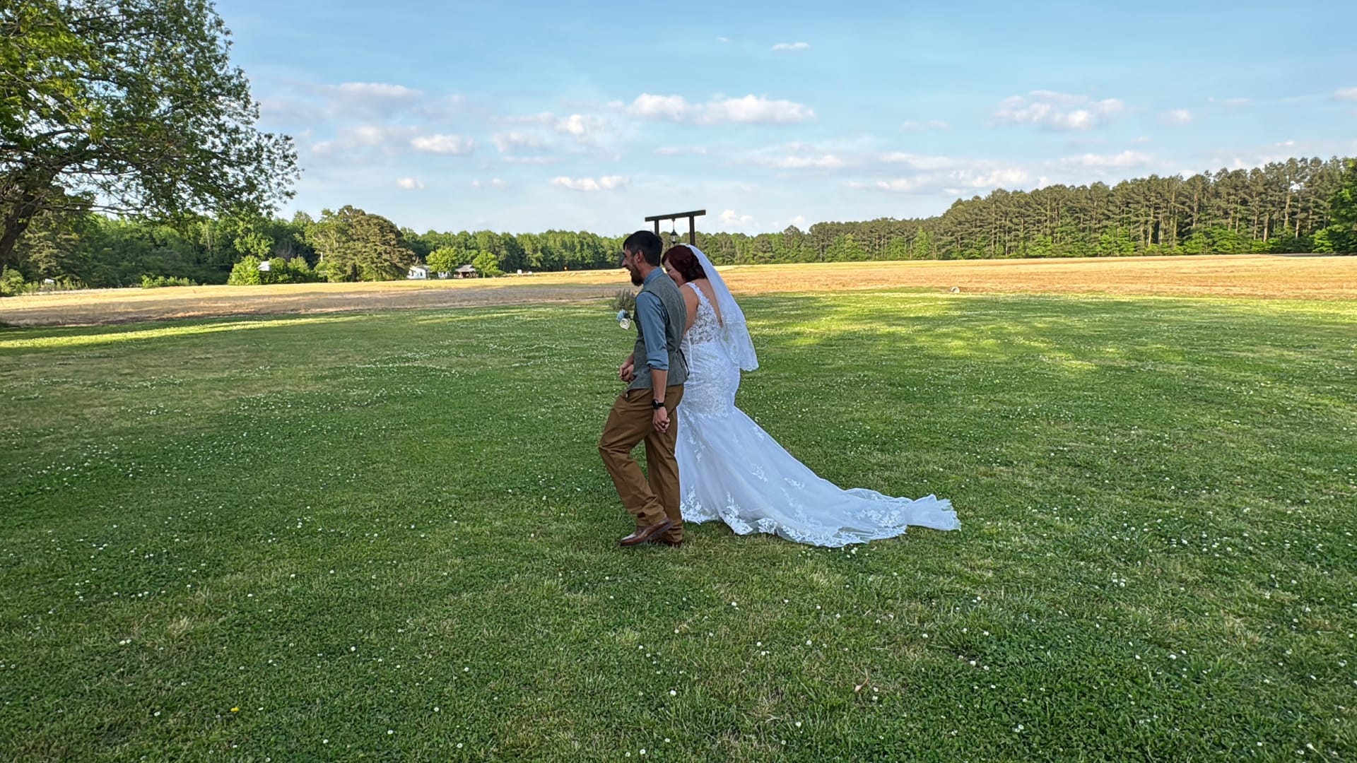 Bride in a lace mermaid gown with a long trailing veil and groom in khaki trousers walk hand in hand across a green lawn with open fields and pine trees at Scarborough House.