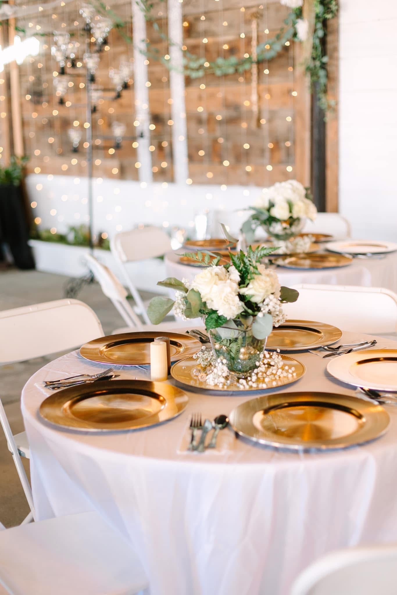 Elegantly set round reception tables with gold charger plates, white hydrangea and eucalyptus centerpieces, and warm string lights in the background at Scarborough House.