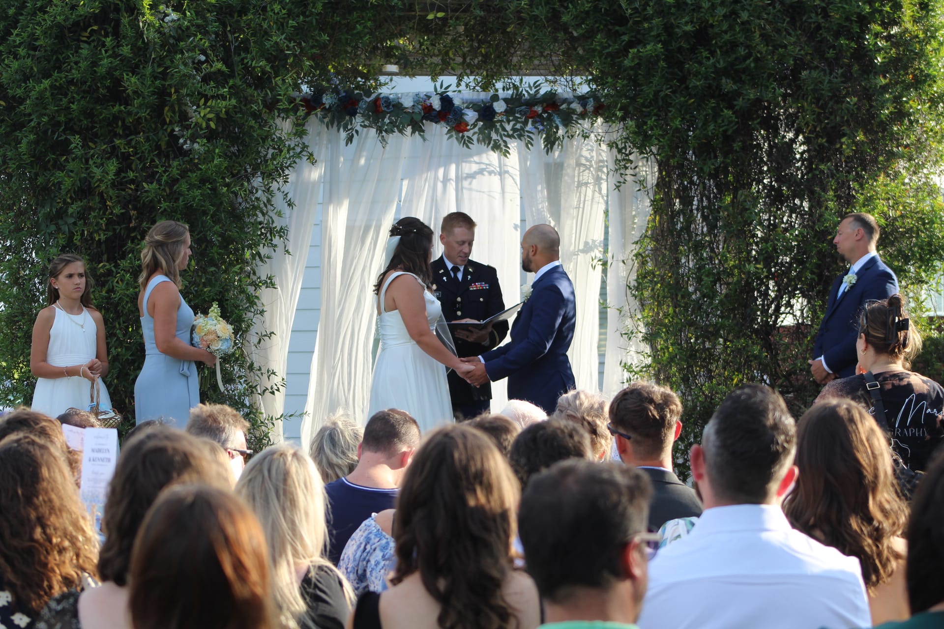 Bride and groom hold hands during outdoor wedding ceremony beneath a floral-adorned draped arch at Scarborough House historic estate, surrounded by lush greenery and seated guests.