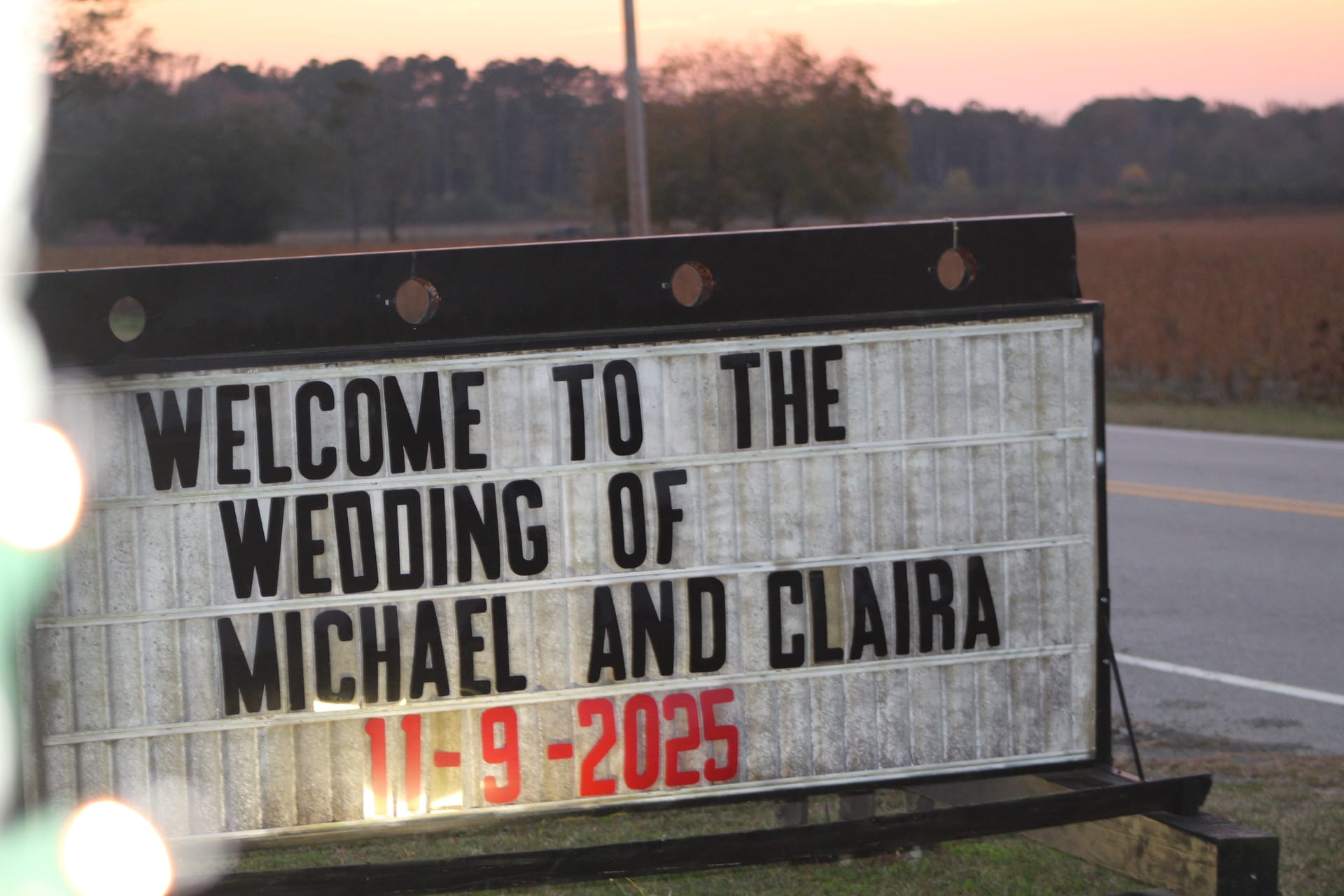 Illuminated roadside marquee sign welcoming guests to the wedding of Michael and Claira on November 9 2025 at dusk with farmland fields and trees in the background.