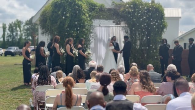 Outdoor wedding ceremony on the Scarborough House lawn with couple exchanging vows beneath a lush greenery and white draped arbor, surrounded by bridesmaids in black and seated guests.