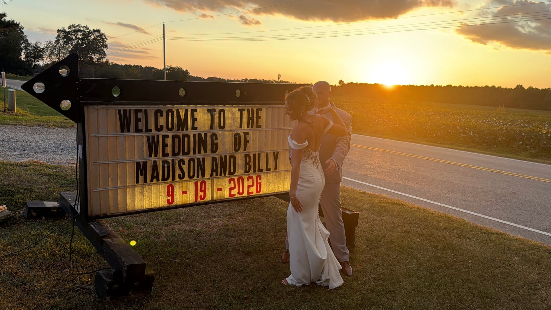 Bride and groom embrace beside a lit marquee welcome sign at golden hour sunset along a rural road at Scarborough House wedding venue.