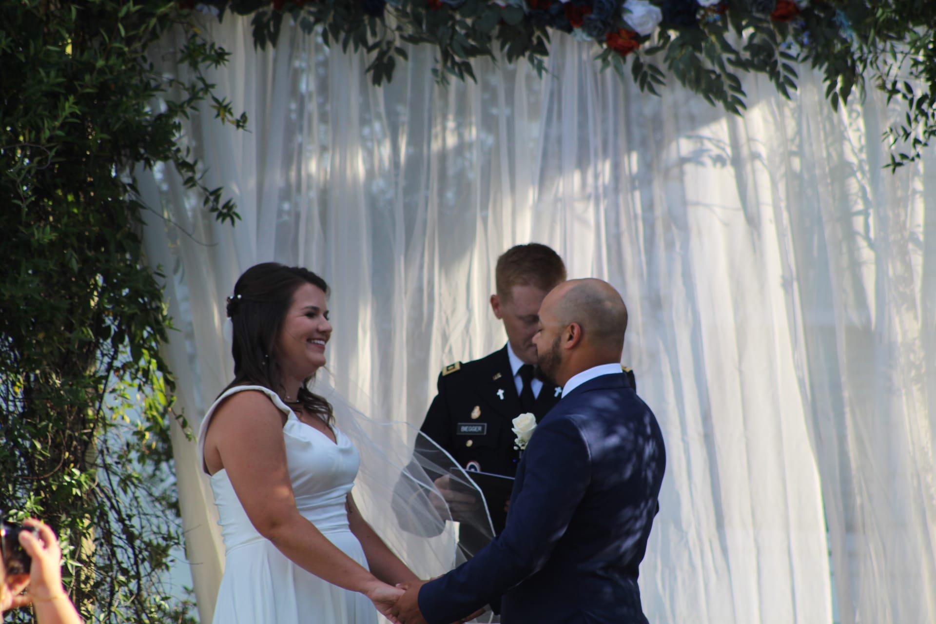Bride in white gown smiling joyfully at groom in navy suit as they hold hands during their outdoor wedding ceremony beneath a floral and white draped arch at Scarborough House.