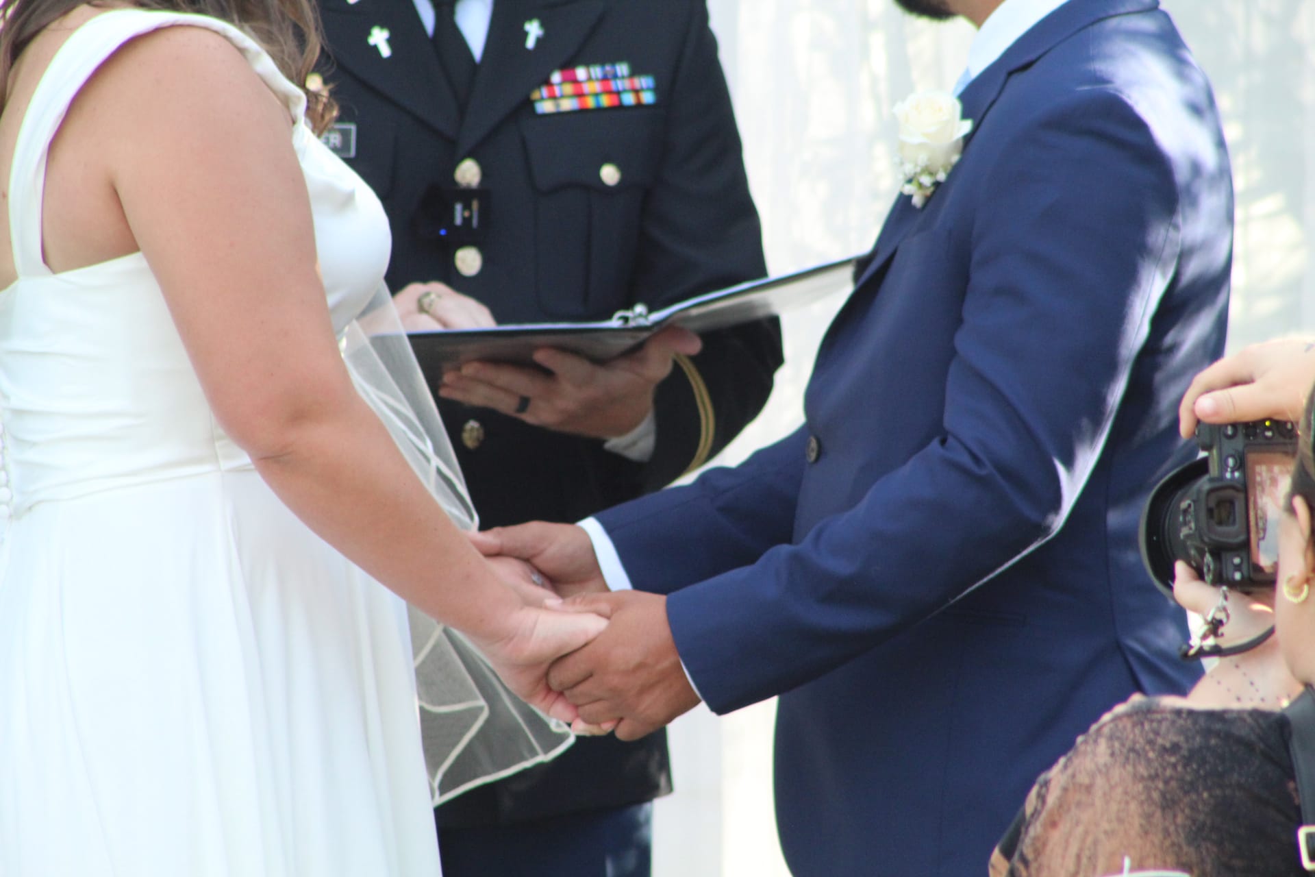 Bride in white gown and groom in navy suit hold hands during wedding vows officiated by a military chaplain at an outdoor ceremony.