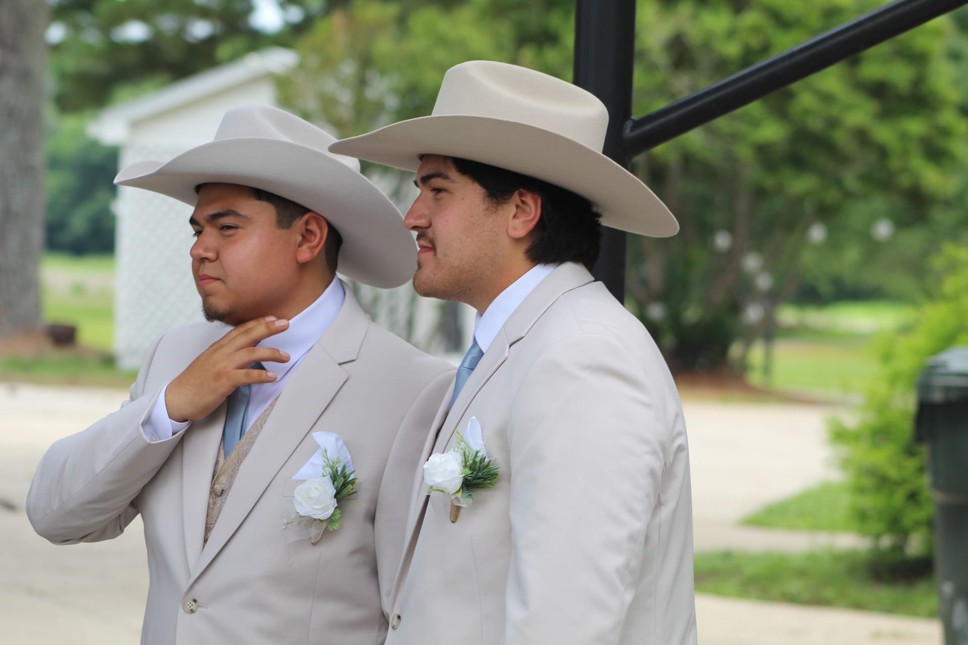 Two groomsmen wearing matching tan cowboy hats and cream suits with white rose boutonnieres stand in profile at an outdoor wedding venue surrounded by lush green trees.