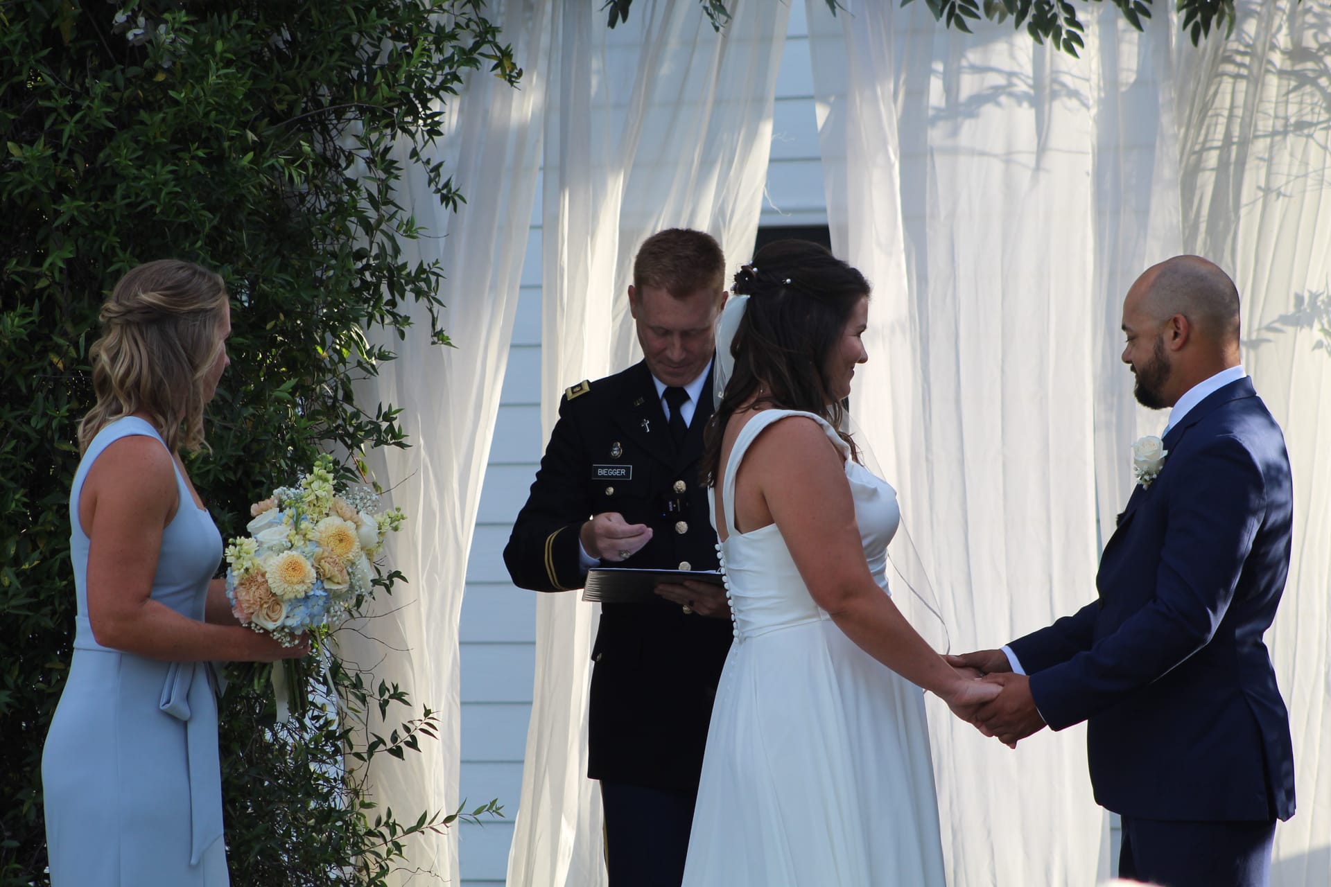 A bride and groom hold hands and smile during their outdoor wedding ceremony officiated by a military officer in dress uniform beneath a white draped arch at Scarborough House.