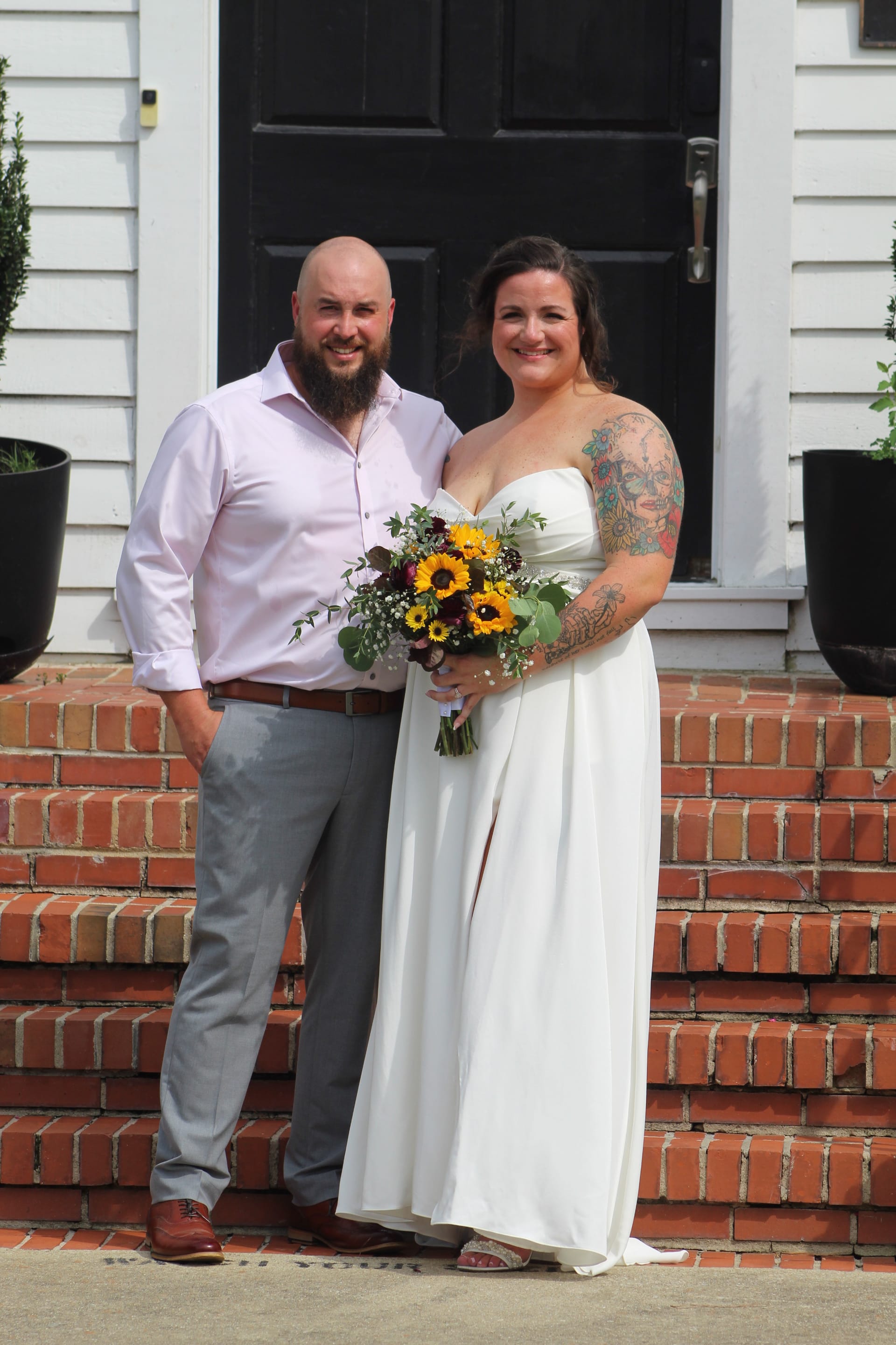 Bride in a white strapless gown holding a sunflower bouquet stands with her bearded groom in a light pink shirt and grey trousers on the brick front steps of the historic Scarborough House estate.