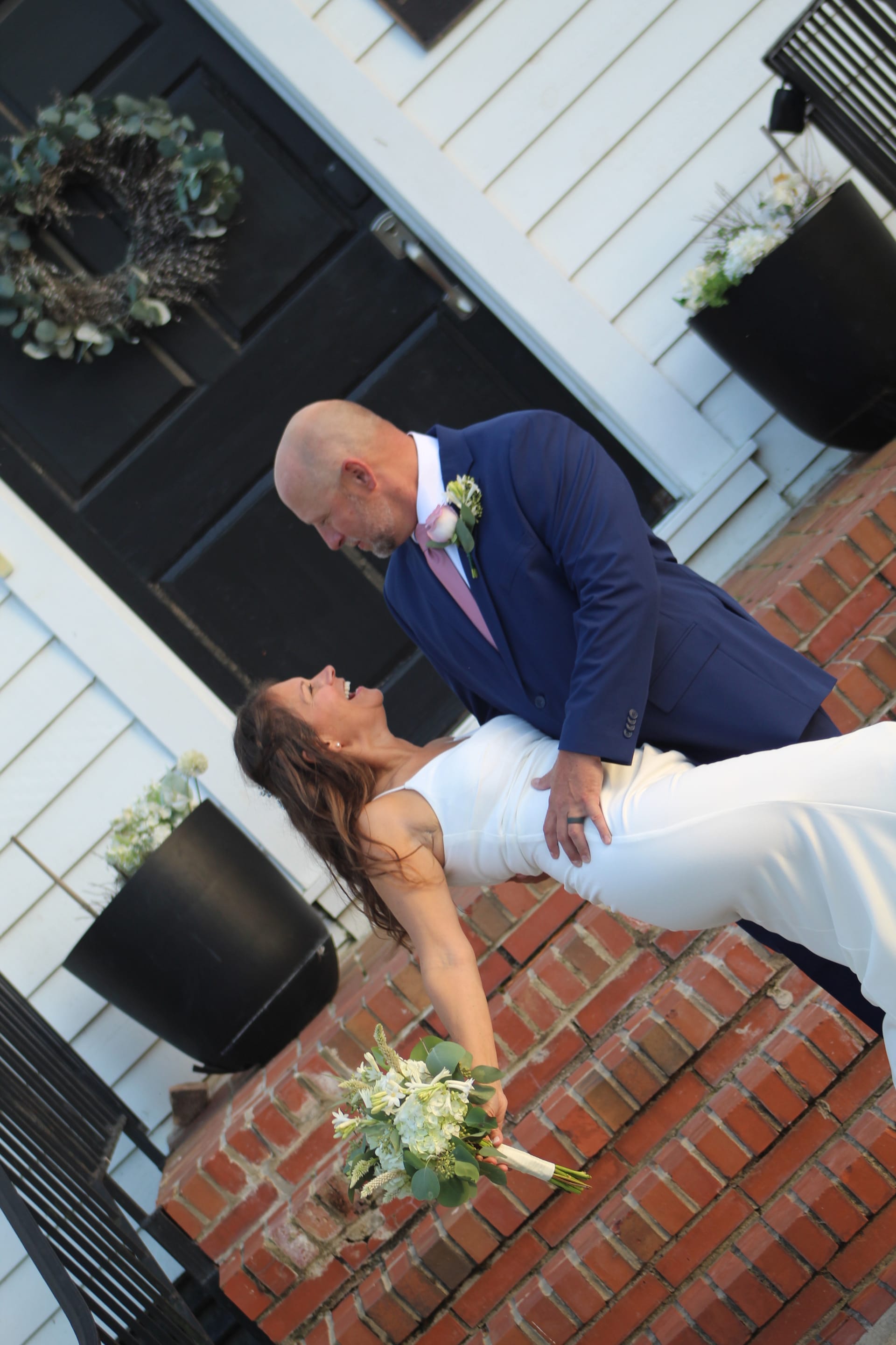 Groom in navy suit dipping his laughing bride in a white gown holding a white floral bouquet on the brick courtyard outside the historic 1821 Scarborough House manor.