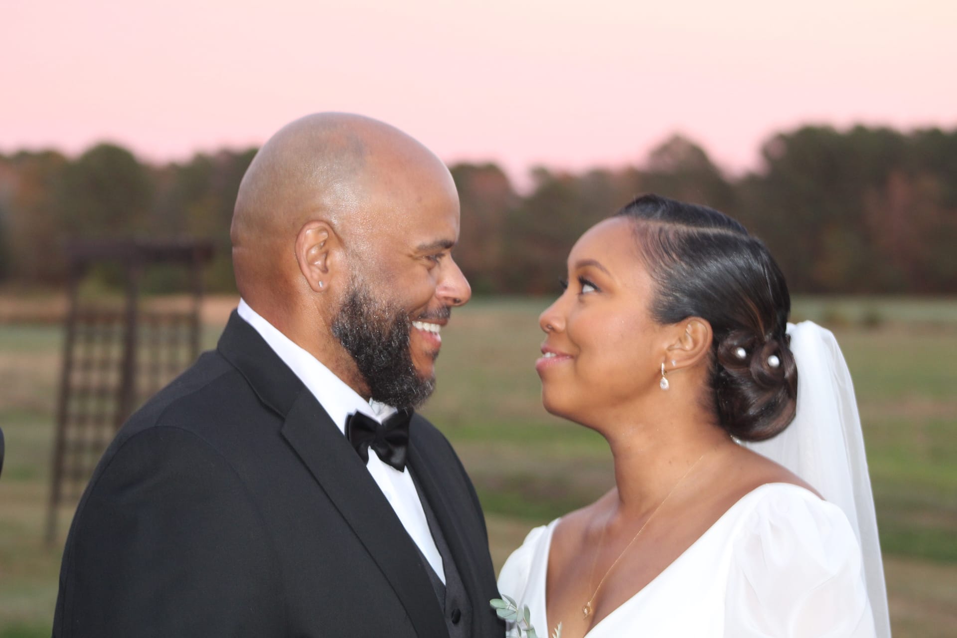 Bride in white gown and groom in black tuxedo gazing at each other and smiling against a pink sunset sky over open farmland fields at Scarborough House.
