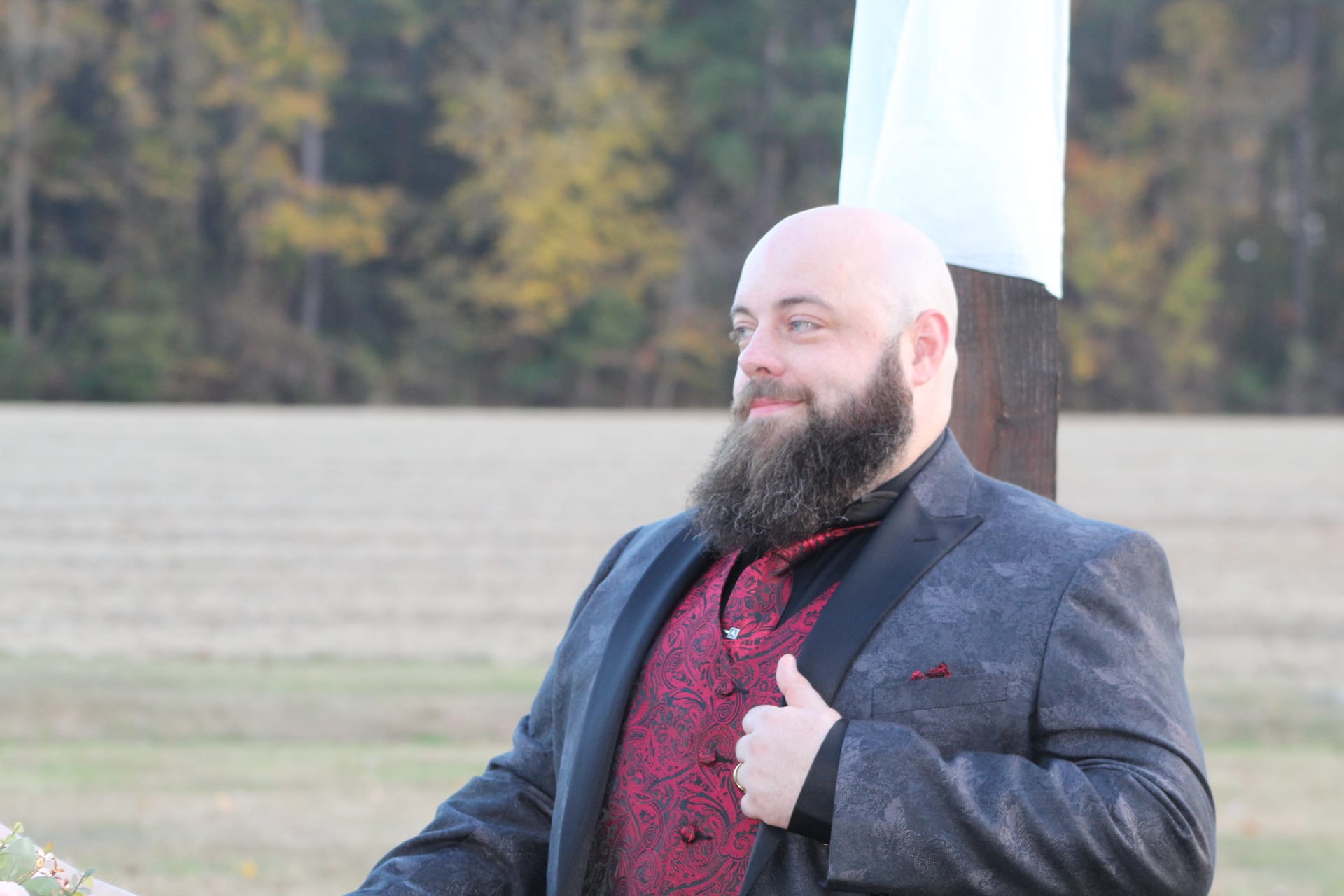 Bald groom with a long beard wearing a burgundy brocade vest and dark tuxedo jacket stands confidently on the ceremony lawn at Scarborough House with autumn trees and open farmland fields in the background.
