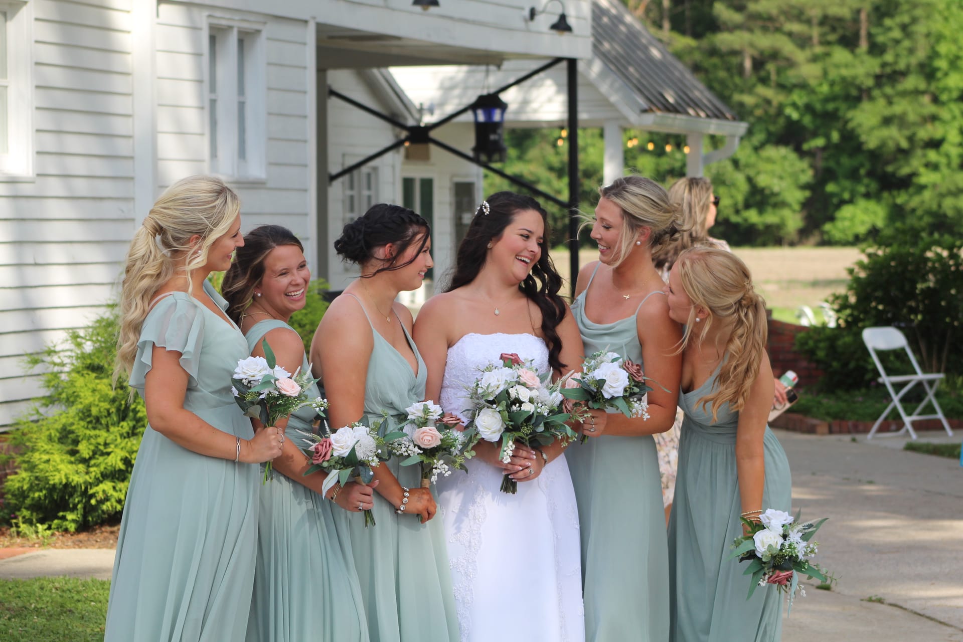 Bride in white strapless gown laughing with five bridesmaids in sage green dresses holding blush and white bouquets outside the historic Scarborough House manor.