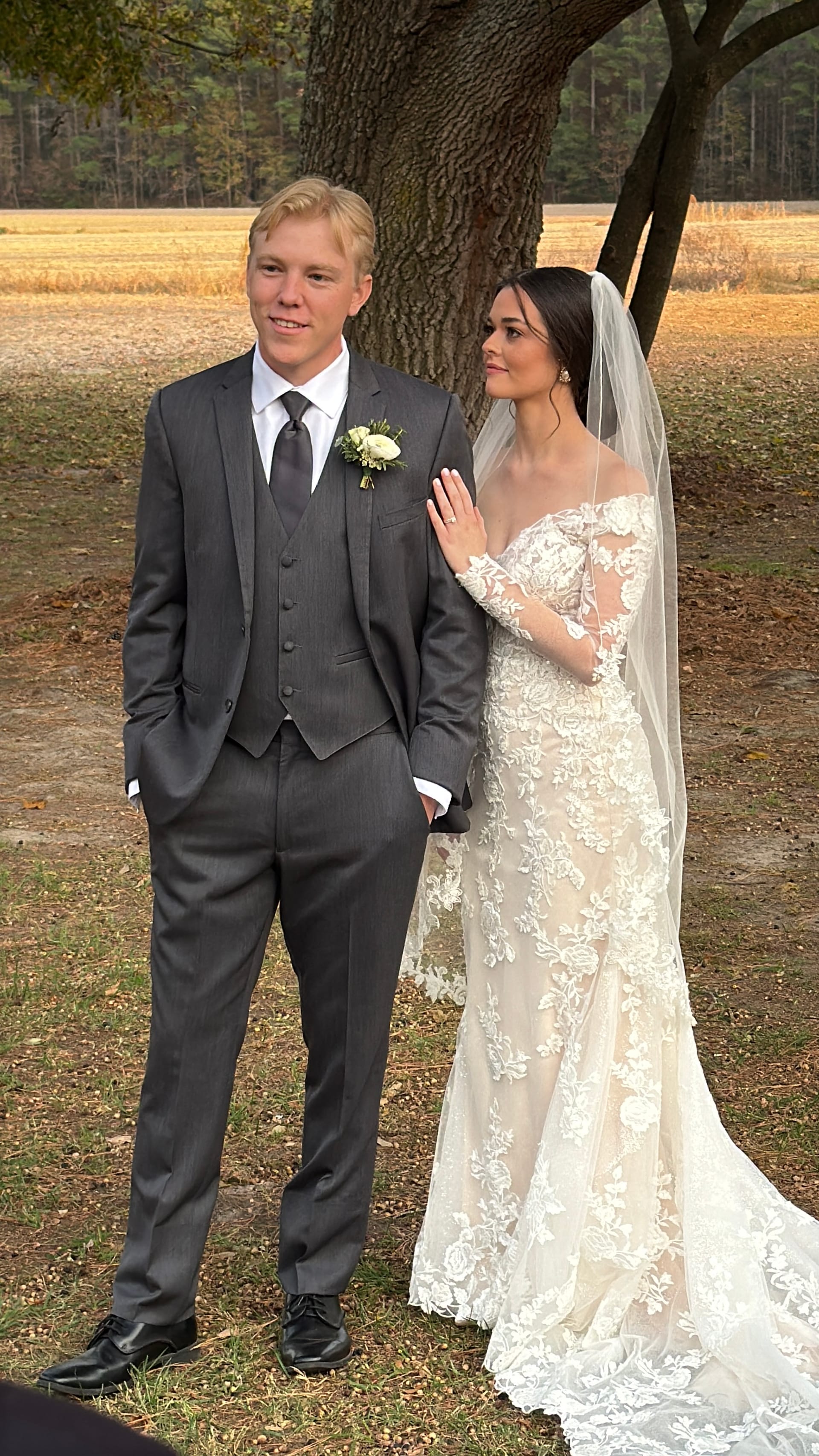 Bride in a long-sleeve lace gown and veil rests her hand on the groom's chest as they stand together beneath a large oak tree with golden fields behind them at Scarborough House.