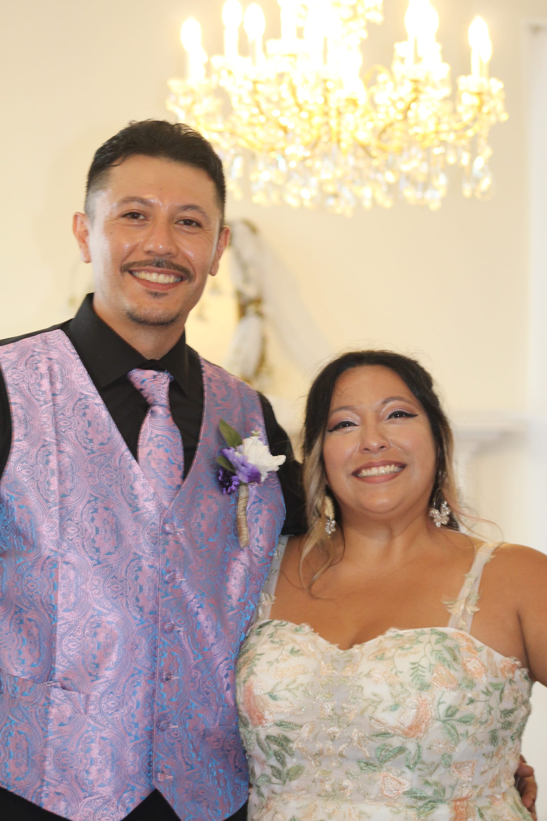 Smiling bride in a floral gown and groom in a purple paisley vest and tie pose together beneath a crystal chandelier inside the grand hall at Scarborough House.