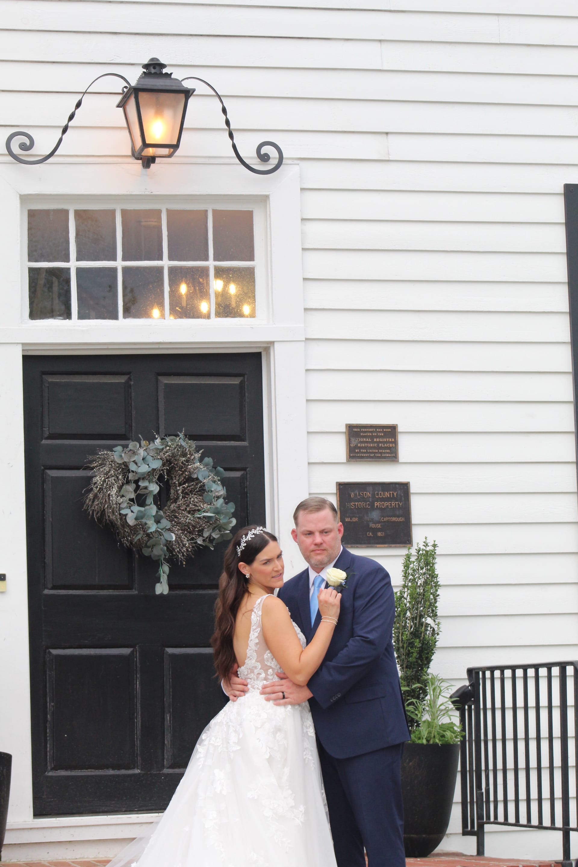 Bride in a lace ballgown and groom in a navy suit embrace in front of the black door and wreath-adorned entrance of the 1821 Scarborough House historic manor.