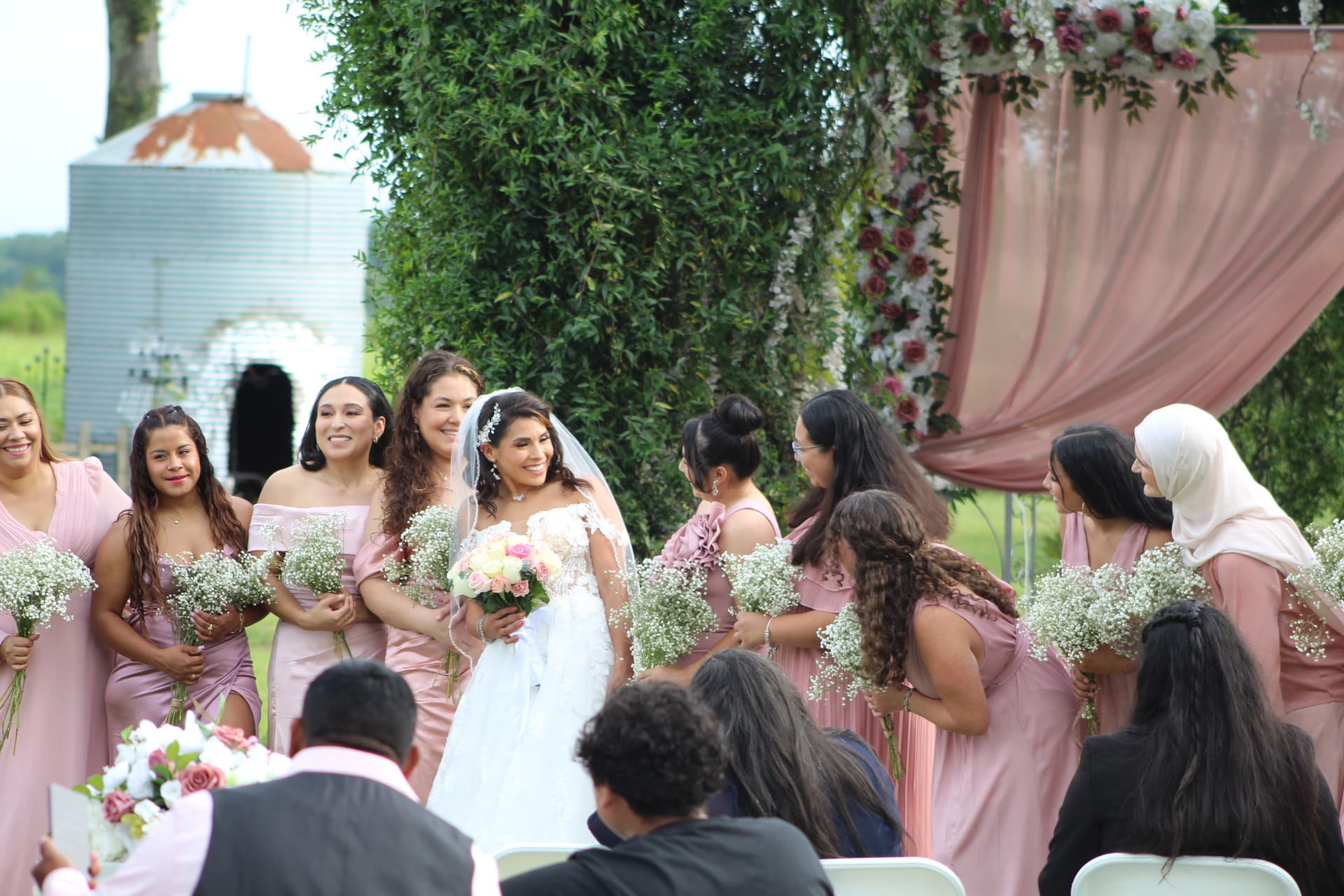 Smiling bride in a white lace gown and veil stands among bridesmaids in blush pink dresses holding baby's breath bouquets during an outdoor ceremony framed by a floral-draped arch and lush greenery.
