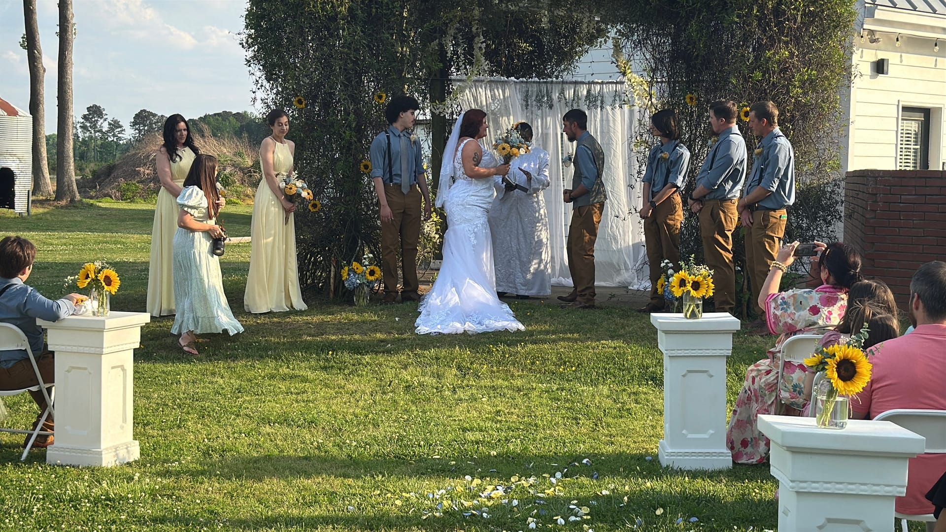 Bride and groom exchange vows beneath a draped floral arch surrounded by bridesmaids in yellow and groomsmen in denim on a sunlit ceremony lawn decorated with sunflowers.
