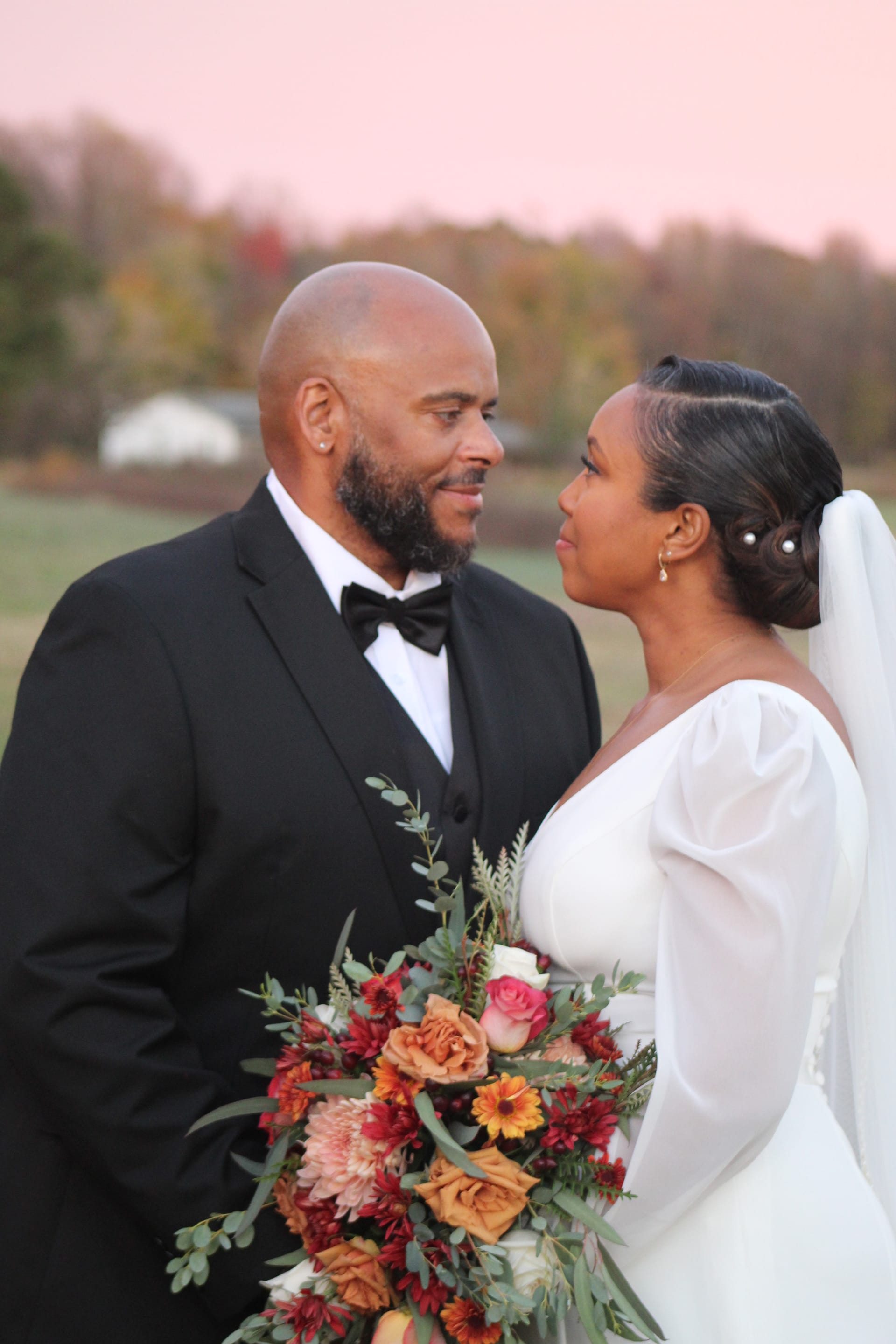 Bride in a white long-sleeve gown and groom in a black tuxedo gaze lovingly at each other during a pink sunset over open farmland fields, bride holding a lush autumn bouquet of burgundy roses and orange dahlias.
