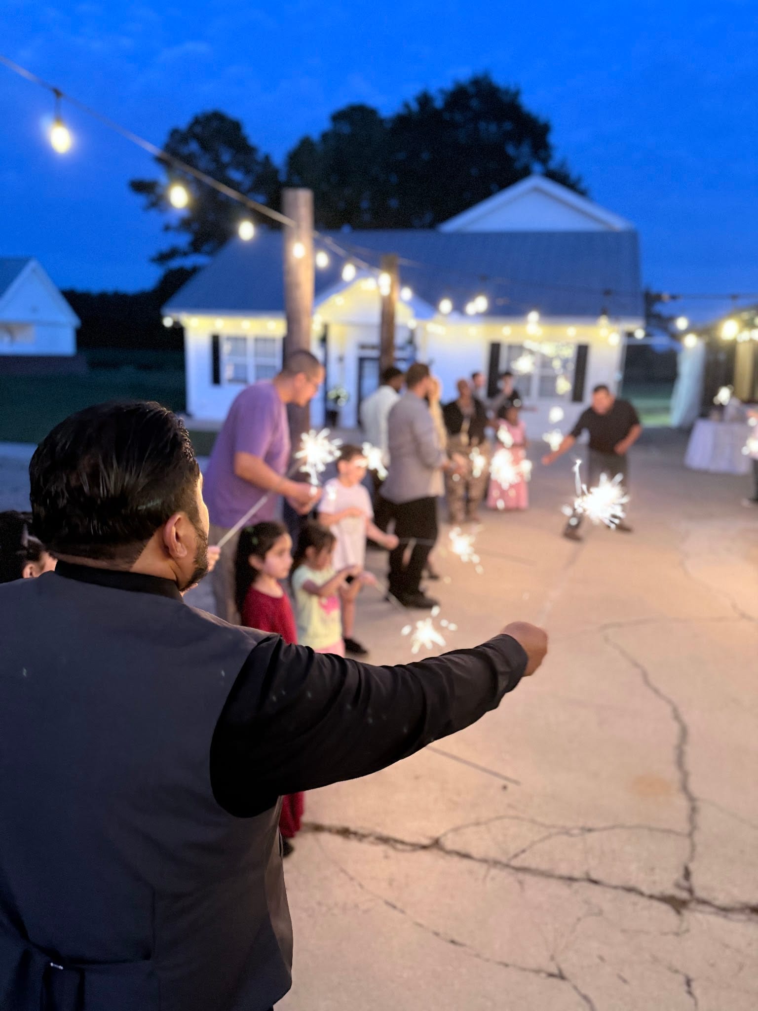 Guests and children line a driveway holding lit sparklers at dusk during a wedding send-off at Scarborough House, with warm string lights glowing above the historic white manor in the background.