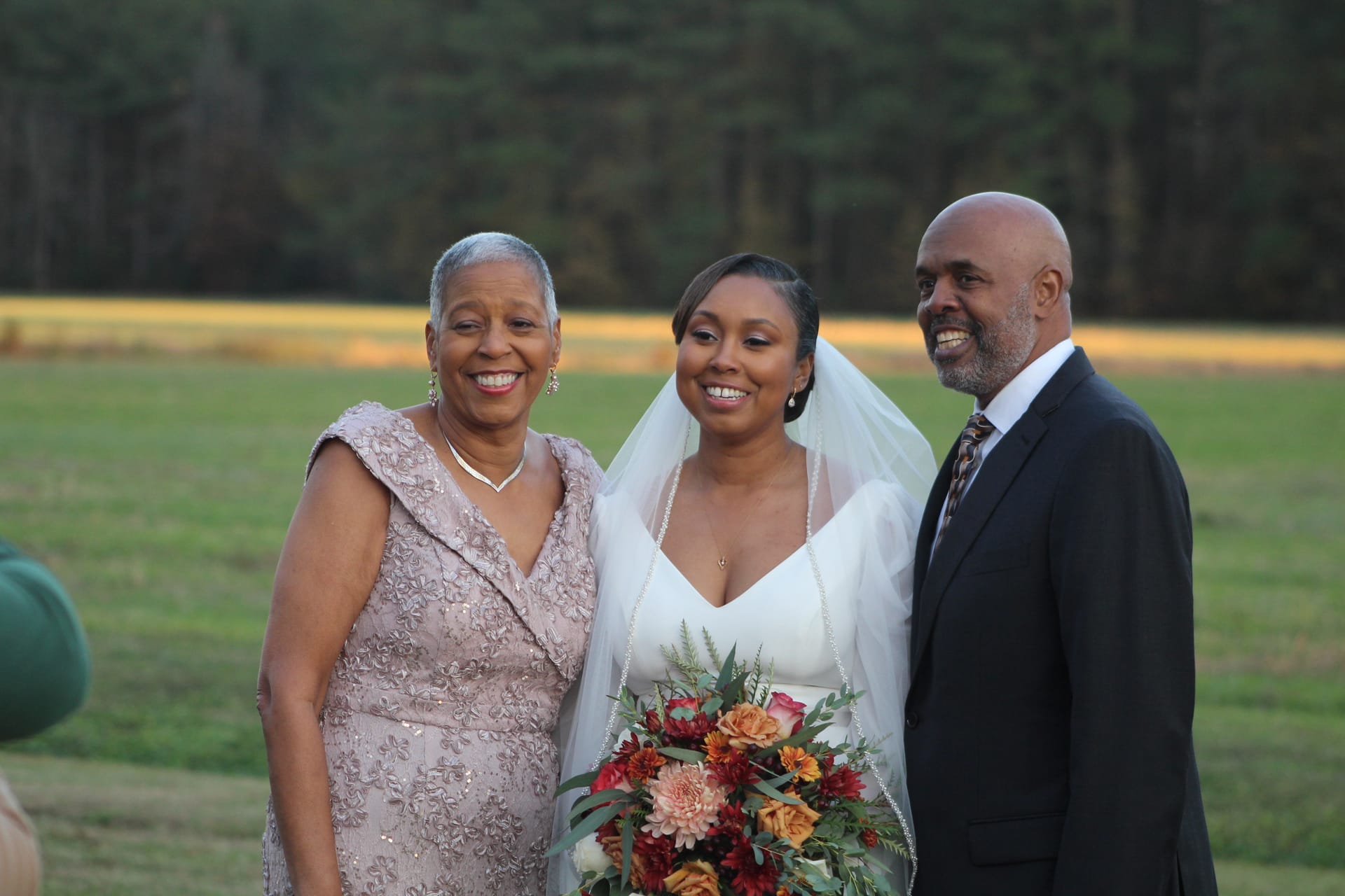 Bride in a white gown and veil holding an autumn bouquet poses and smiles with her mother in a blush gown and her father in a dark suit on the open farmland fields of a historic estate wedding venue at golden hour.