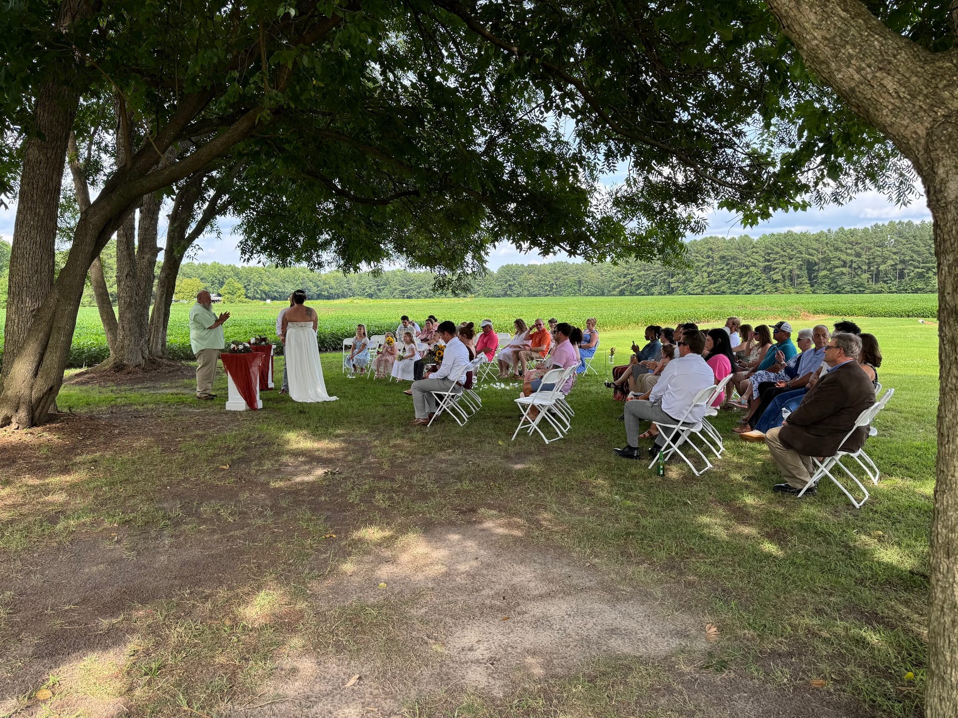 An intimate outdoor wedding ceremony held under large oak trees on the ceremony lawn at Scarborough House, with guests seated in white folding chairs and lush farmland fields in the background.