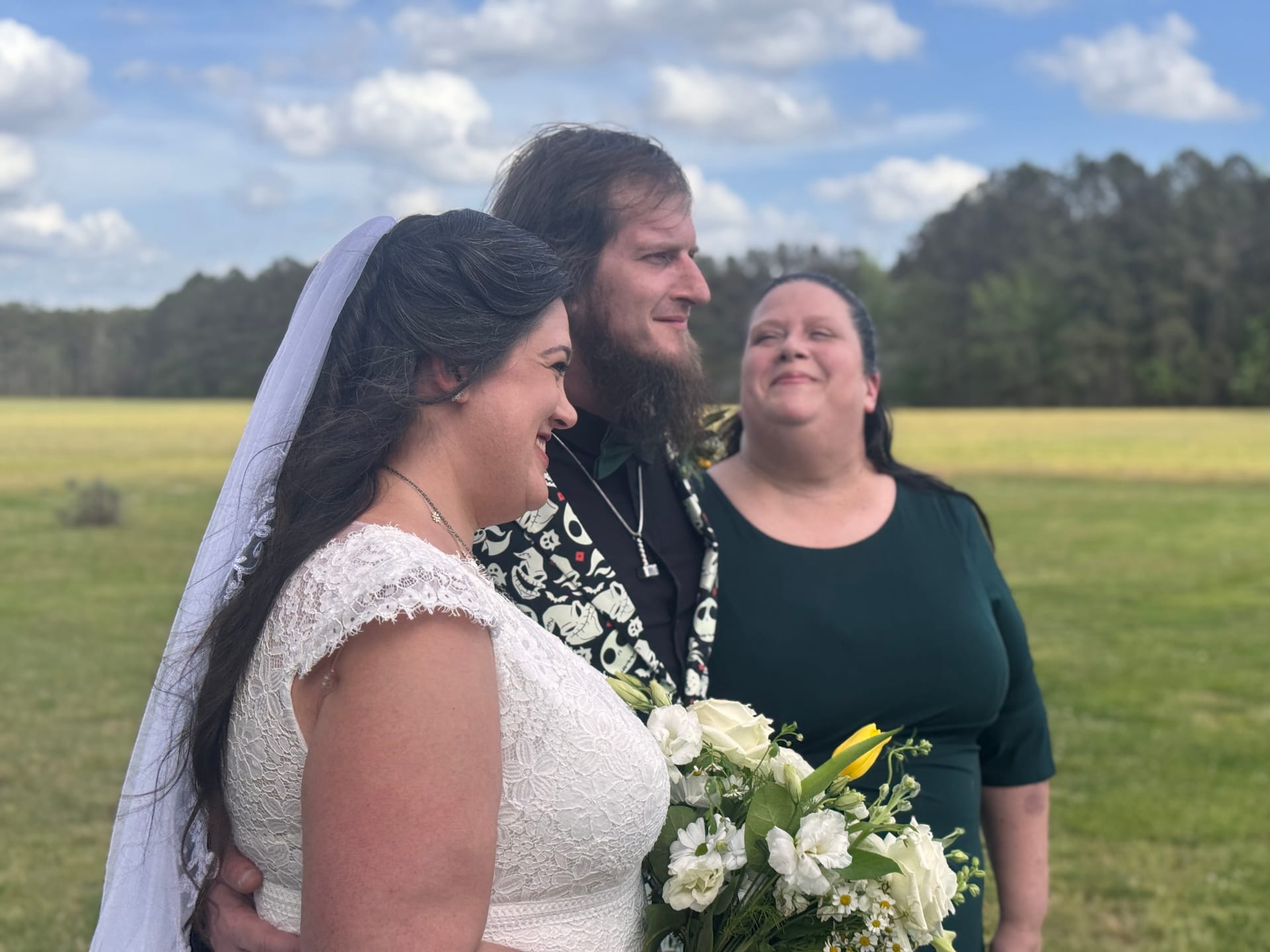 A smiling bride in a white lace gown holding a white and yellow bouquet stands beside her bearded groom during an outdoor ceremony at Scarborough House, with open farmland fields and a blue sky in the background.