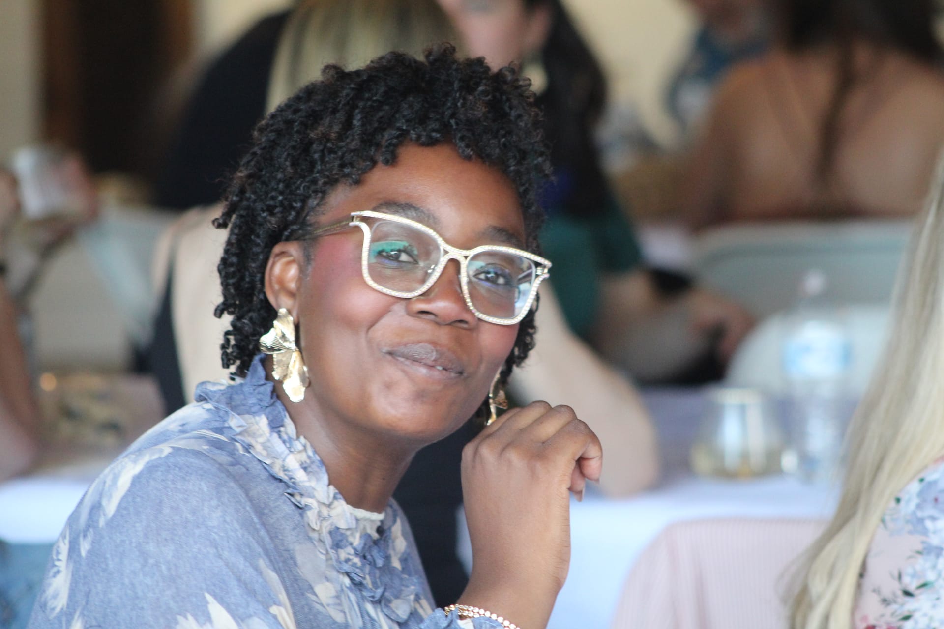 A smiling woman wearing white-framed glasses and gold leaf earrings rests her chin on her hand during a reception at Scarborough House.