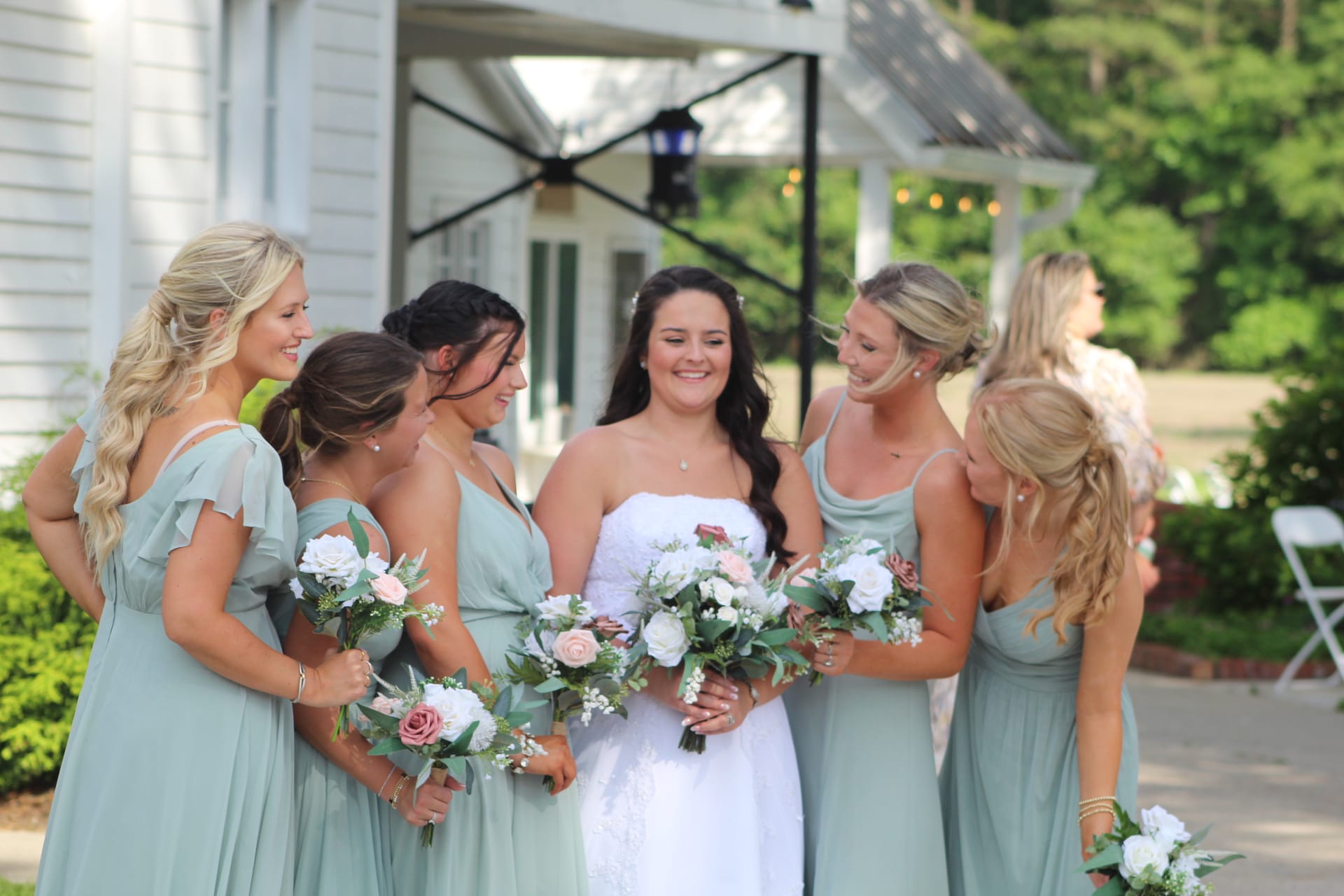 Bride in white strapless gown smiling with five bridesmaids in sage green dresses holding blush and white bouquets outside the historic Scarborough House manor.