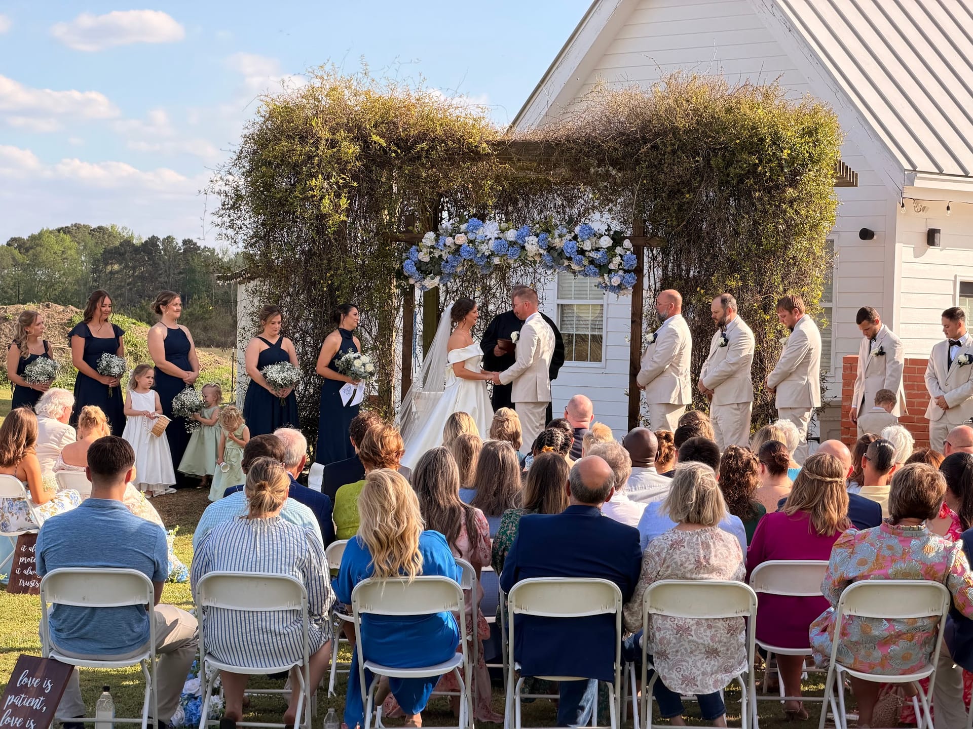 Bride and groom exchange vows beneath a lush greenery and blue floral arch on the ceremony lawn at Scarborough House historic estate, surrounded by bridesmaids, groomsmen, and seated guests.