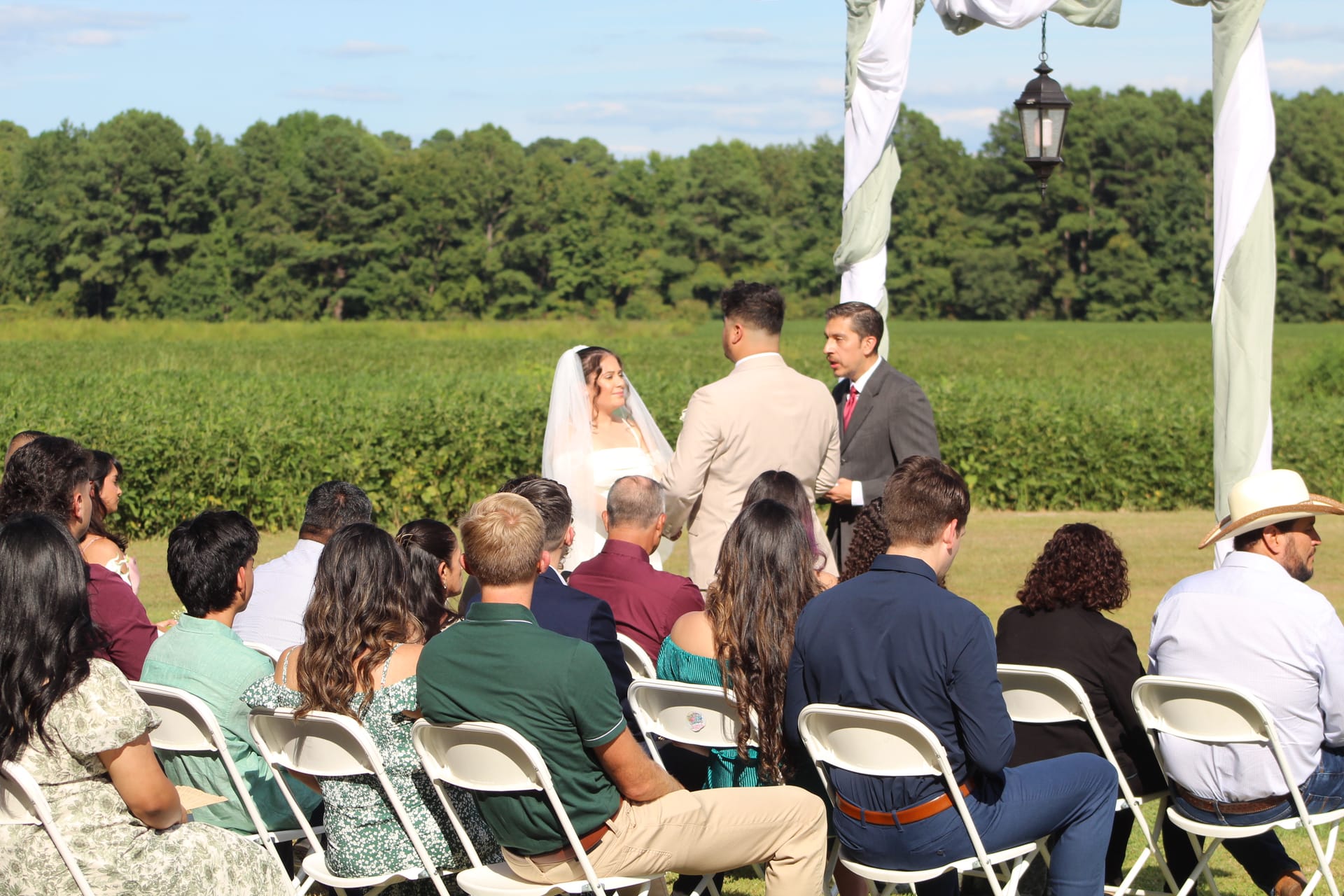 Bride and groom exchange vows beneath a draped arch adorned with a lantern on the ceremony lawn at Scarborough House, surrounded by seated guests with a sweeping green field and tree line in the background.