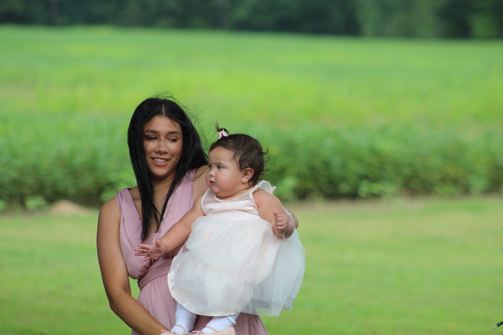 A smiling bridesmaid in a dusty rose gown holds a chubby baby girl dressed in a white tutu on the lush green fields of Scarborough House estate.