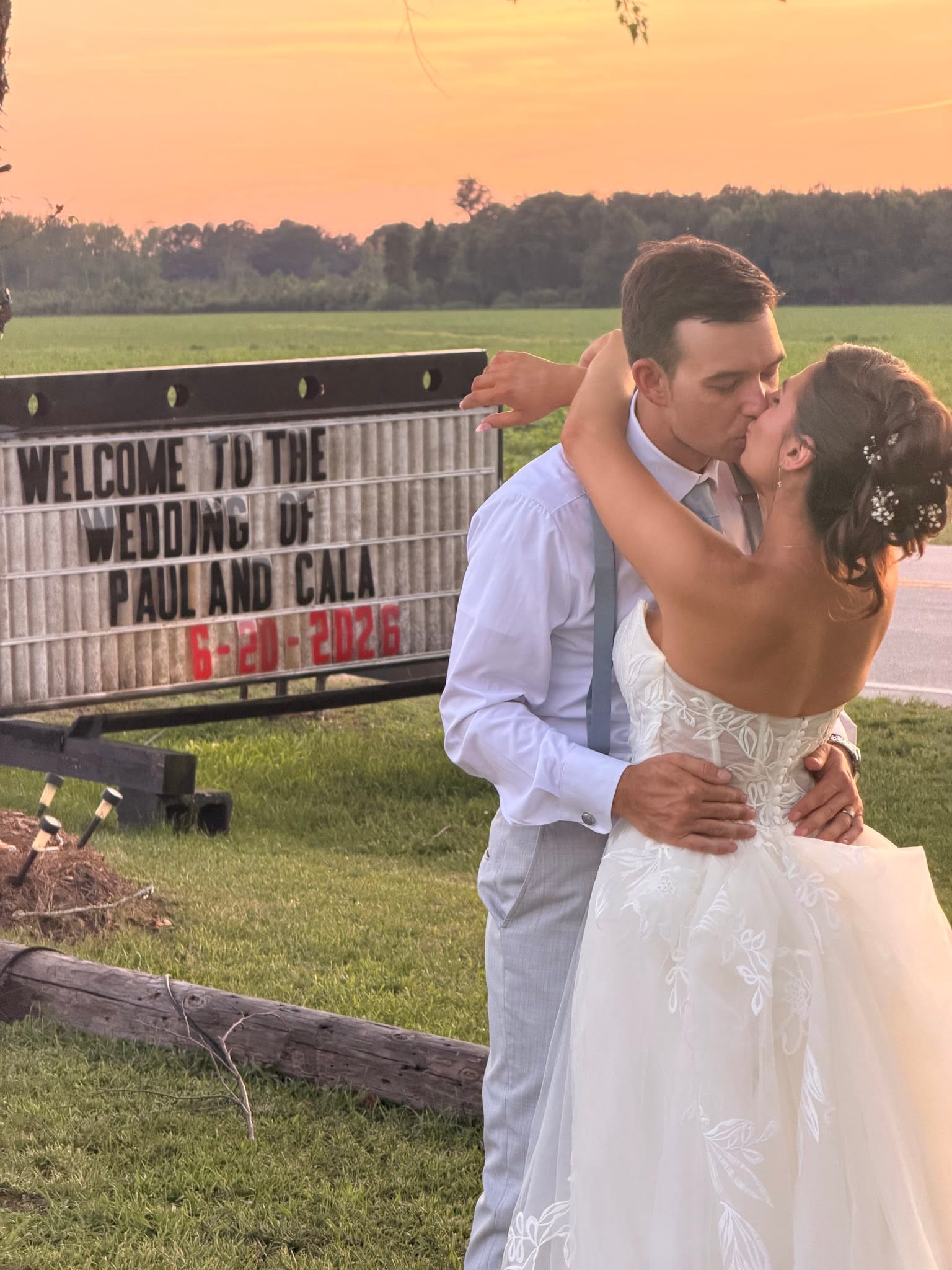 Bride and groom share a kiss in front of a welcome wedding marquee sign at golden-hour sunset with open farmland fields in the background.