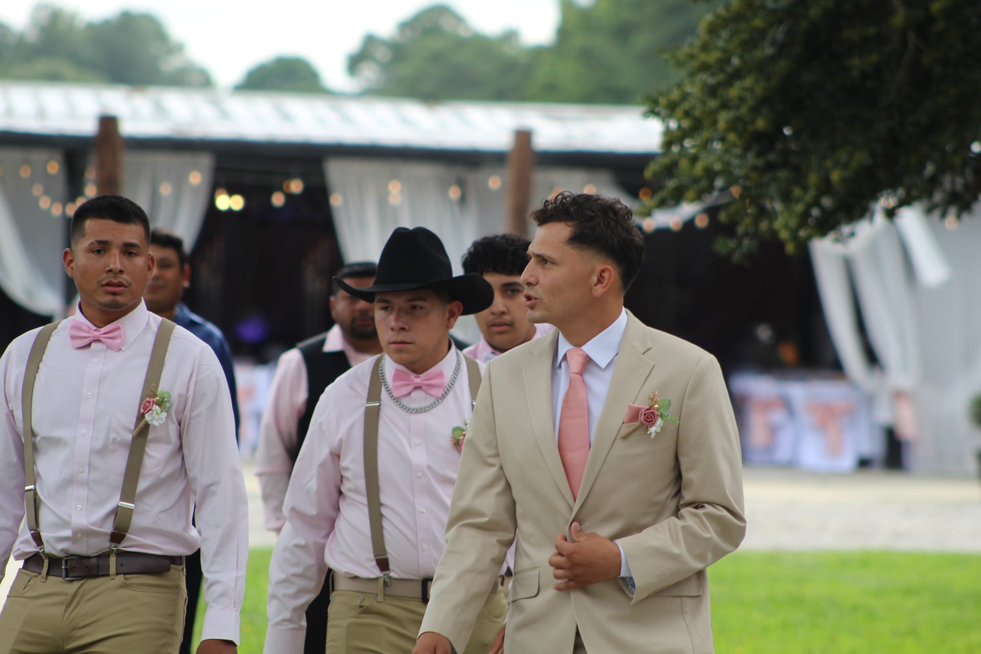 Groom in a tan suit with a pink tie leads his groomsmen in pink shirts, suspenders, and a black cowboy hat across the ceremony lawn at Scarborough House wedding venue.